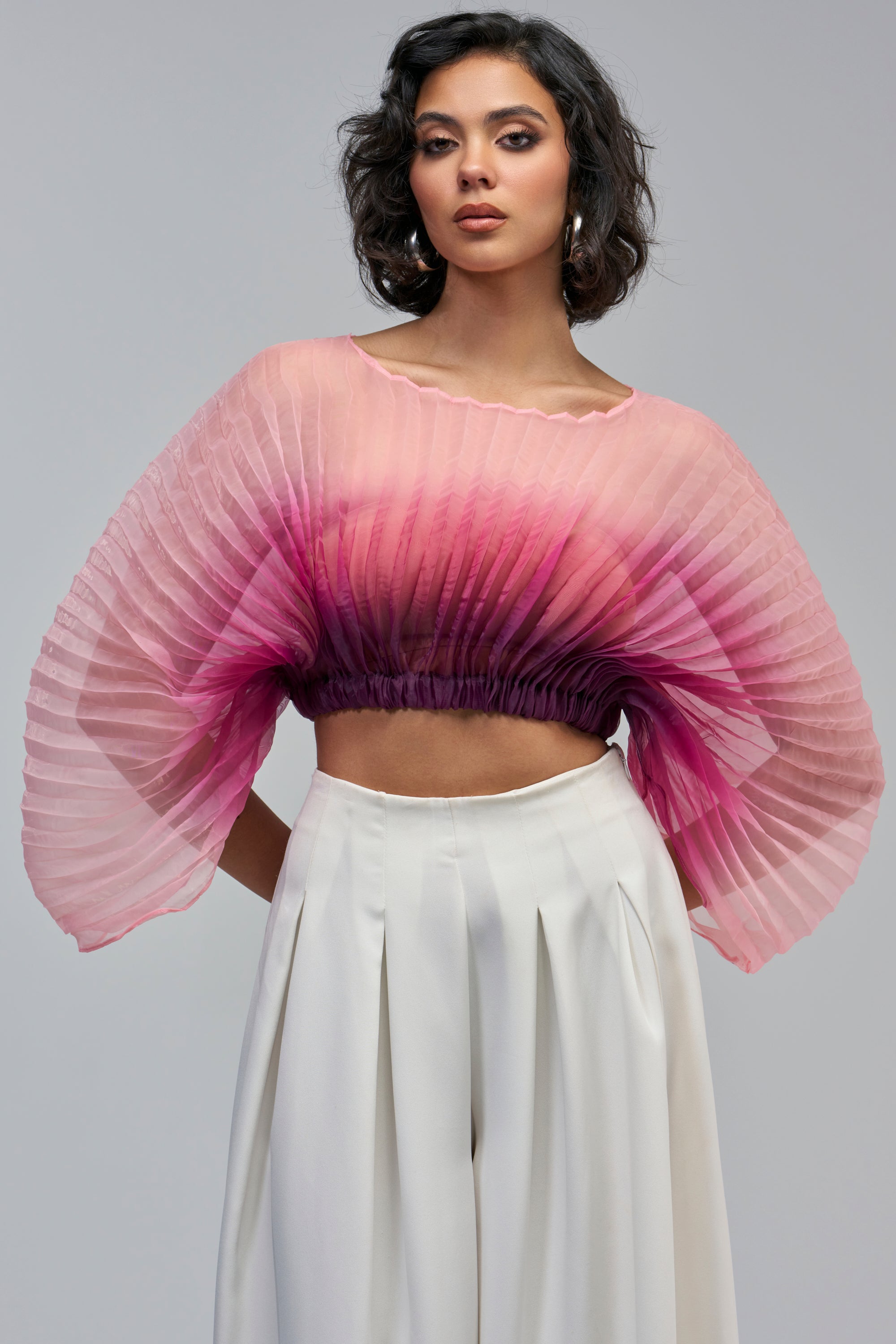 A person wears a PRETTY IN PINK BLOUSE featuring a pink ombre pleated fan design that transitions from light pink at the neckline to deep purple at the waist, with wide, flowing sleeves.