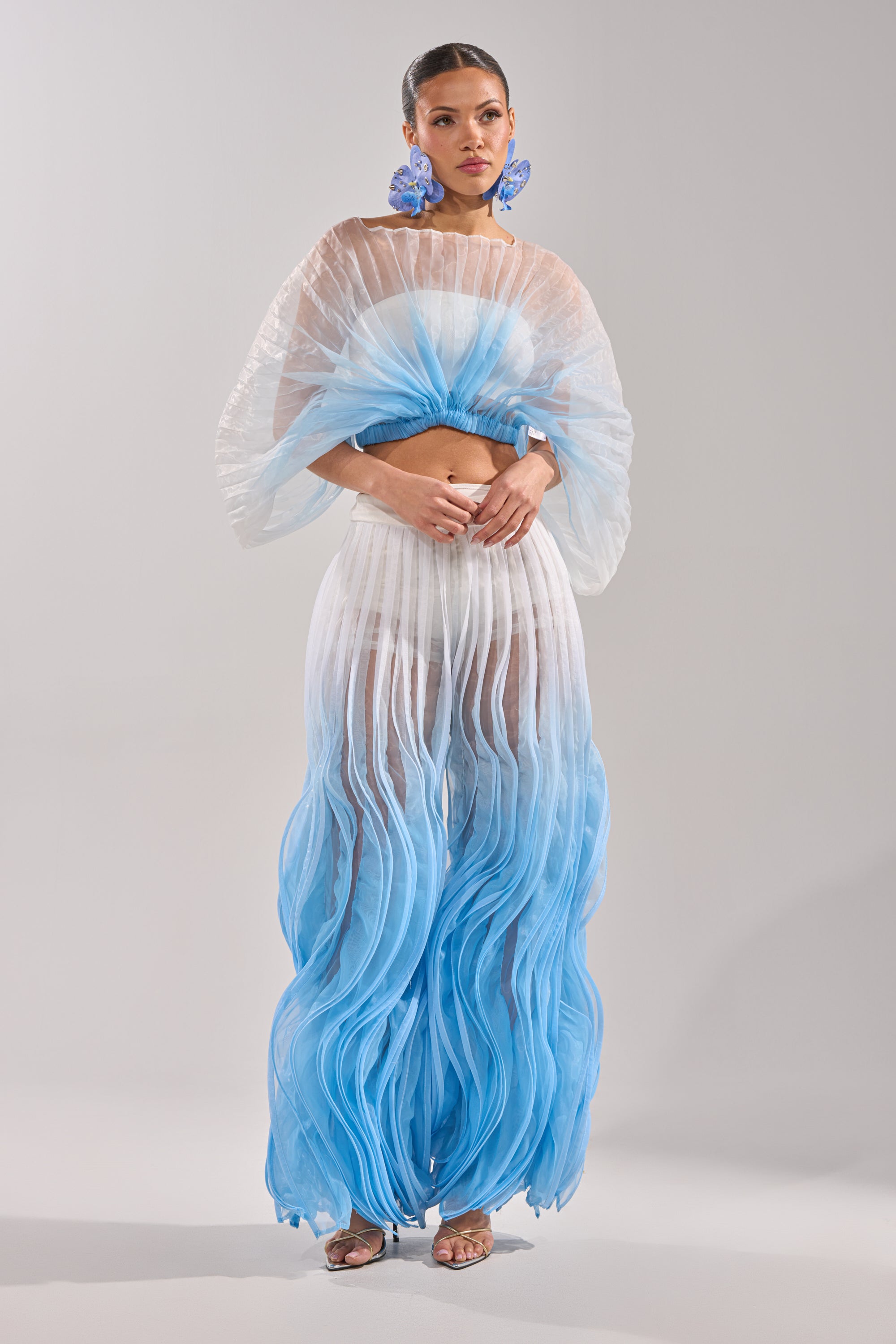 A woman models the IN THE SKY PALAZZO PANT—sheer ombre, wide-leg pants fading from white to blue with pleated, wavy textures—paired with large blue floral earrings. 0