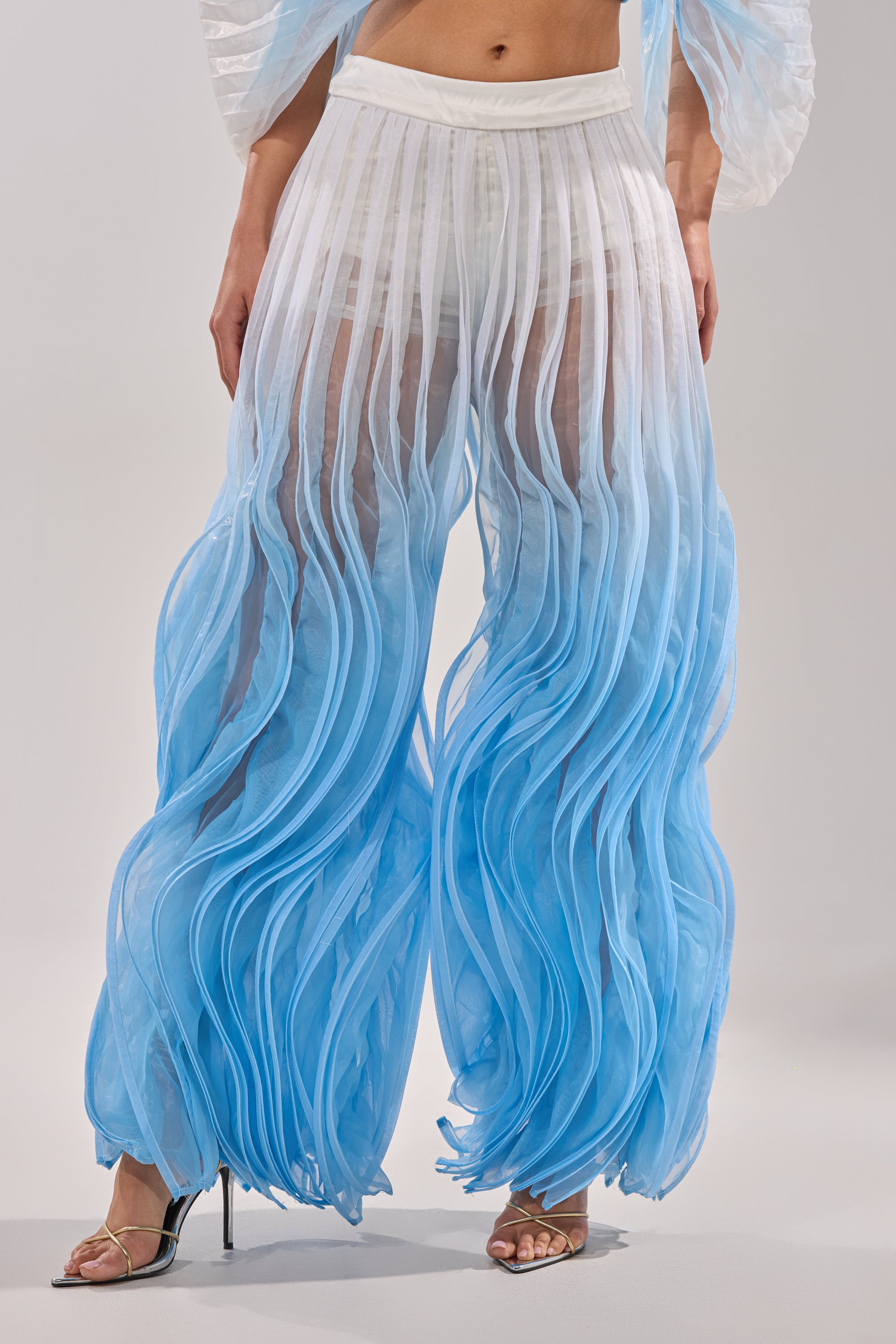 A person wearing the IN THE SKY PALAZZO PANT, featuring high-waisted, sheer ombre pleated wide legs and sculptural wavy details, styled with heeled sandals for an airy, flowing look. 1
