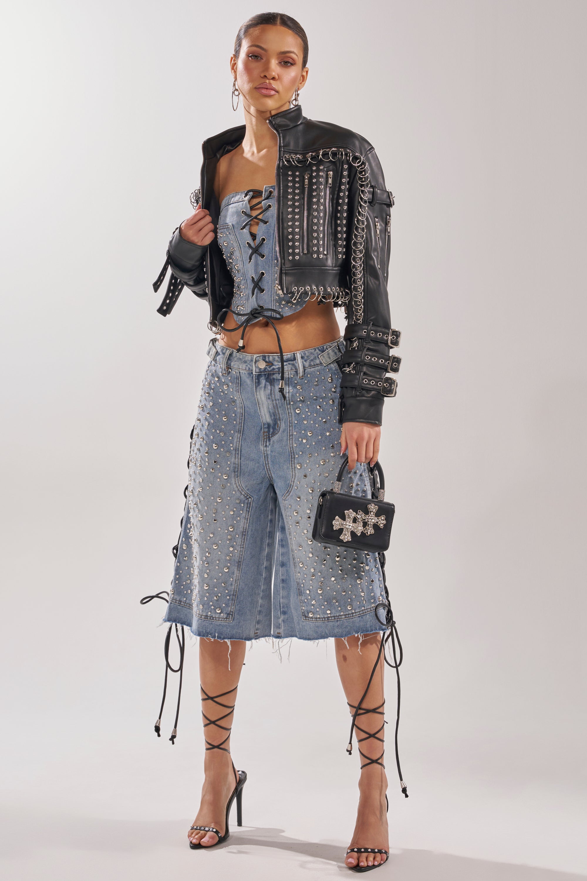 A woman poses against a plain background in a studded leather jacket, lace-up denim bustier, and HONEY BABY BERMUDA SHORT featuring Y2K streetwear details. She carries a black bag and wears strappy high heels. 9