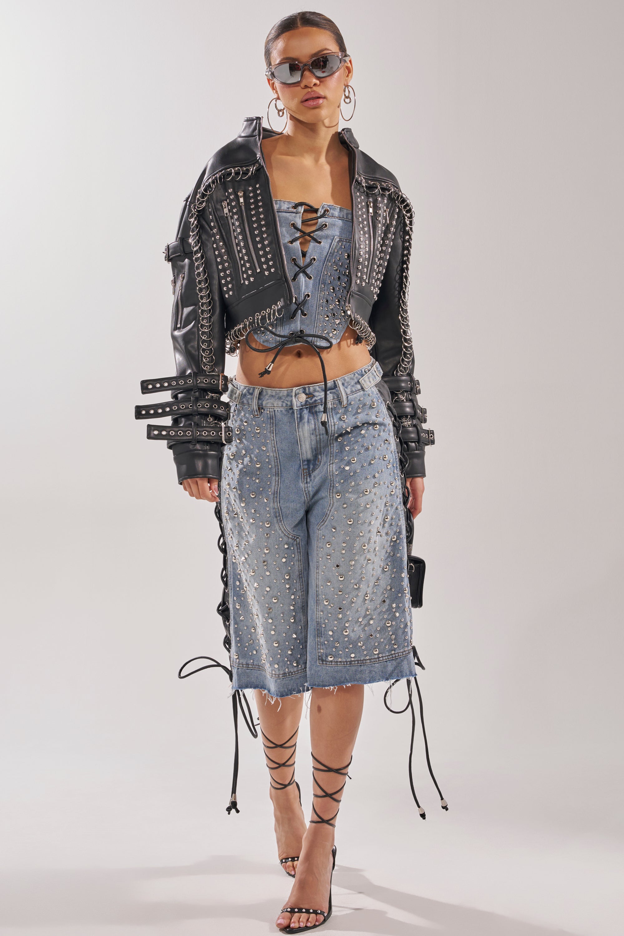 A woman in Y2K streetwear stands against a plain background, wearing sunglasses, a studded leather jacket, a lace-up denim corset top, HONEY BABY BERMUDA SHORT, lace-up high-heeled sandals, and large hoop earrings. 11