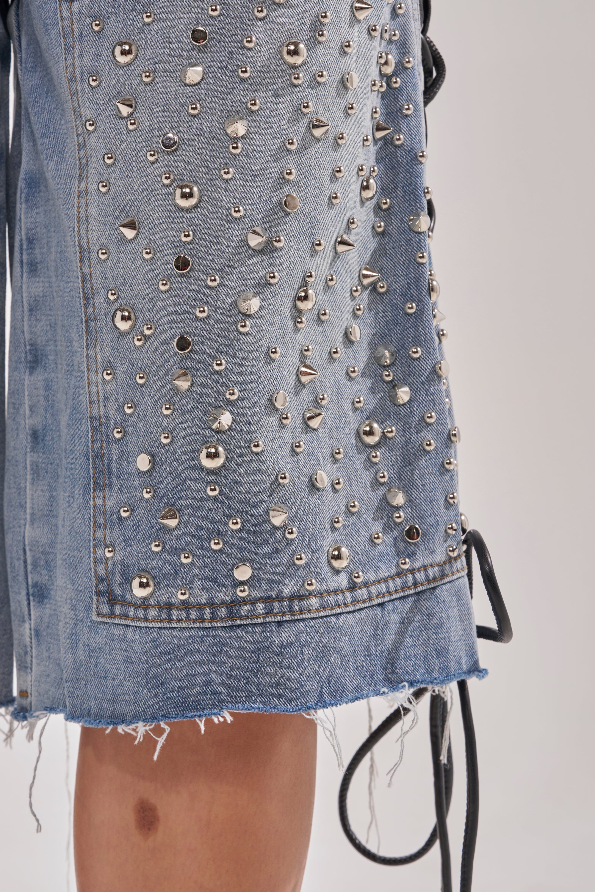 Close-up of the HONEY BABY BERMUDA SHORT: denim lace-up shorts adorned with silver studs and spikes, frayed hem, and black side laces evoke bold Y2K streetwear. A bare leg is partially visible below. 12