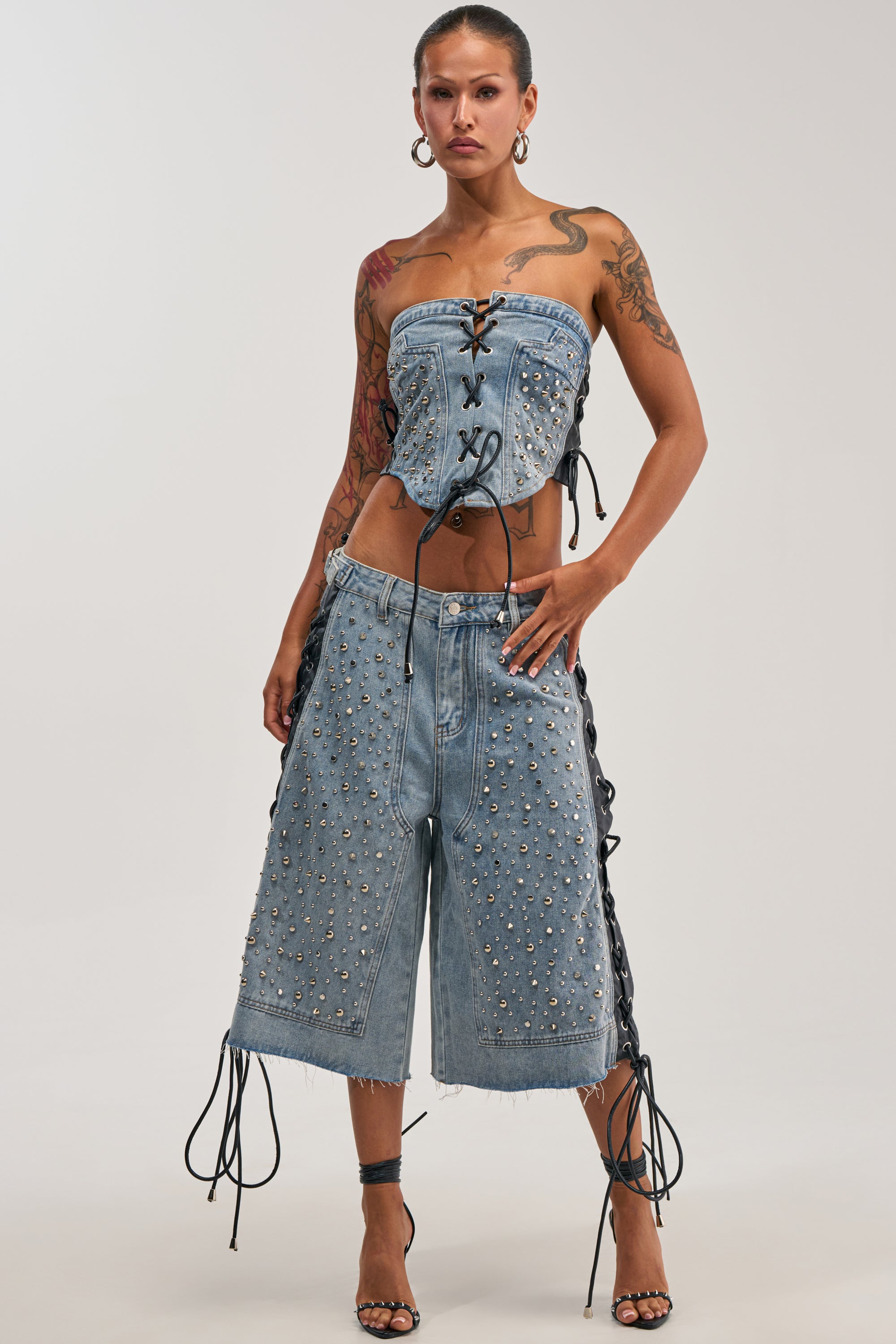 A woman strikes a confident pose in the HONEY BABY BERMUDA SHORT, pairing the studded denim bermuda shorts with a strapless, lace-up denim top featuring edgy lace-up side panels for a bold Y2K streetwear look. She accessorizes with hoop earrings, strappy heels, and displays tattoos on her arms and shoulders.