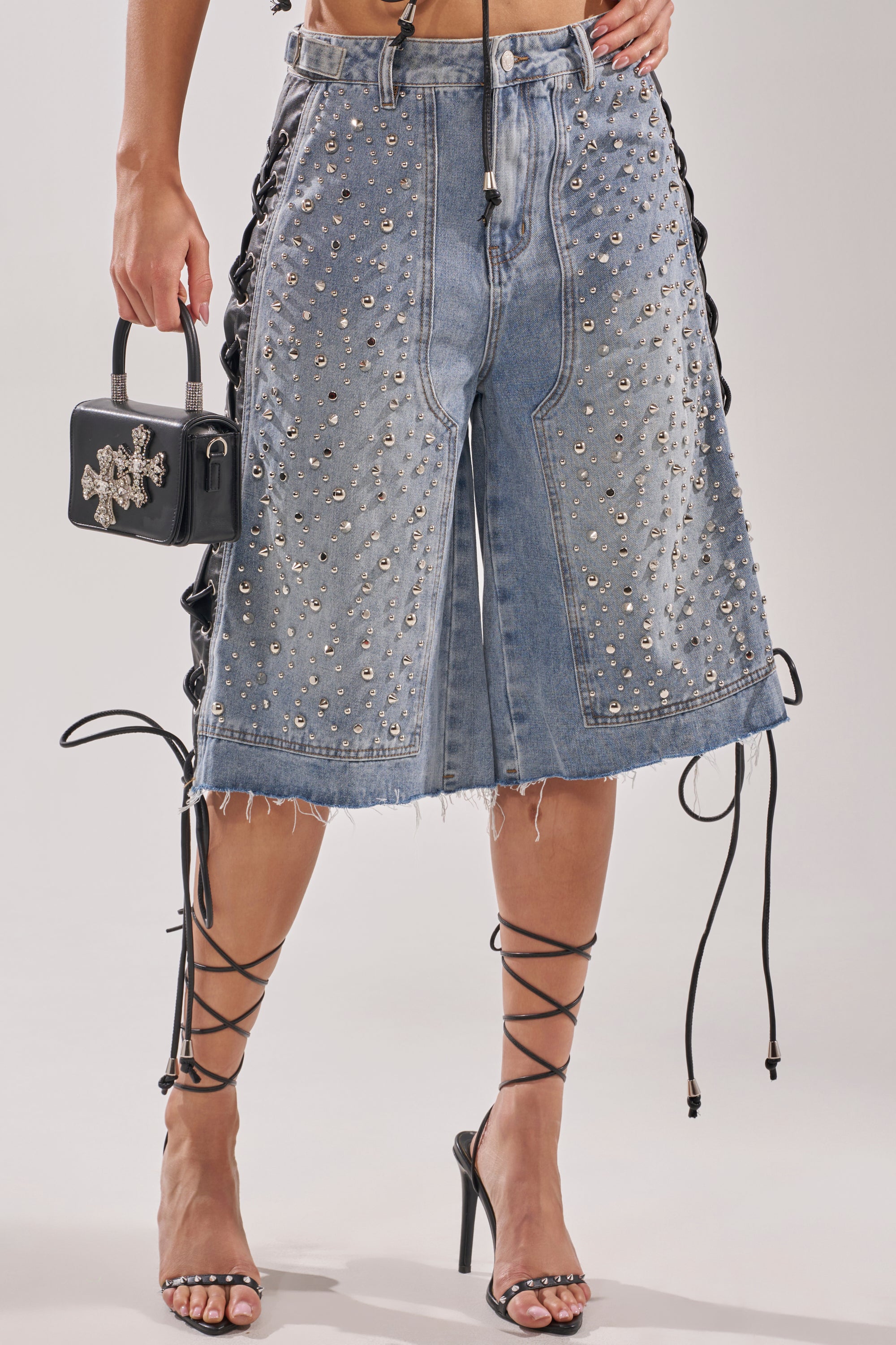 Wearing the HONEY BABY BERMUDA SHORT with studded details, lace-up sides, and frayed hems, she completes this bold Y2K streetwear look with a small black handbag and strappy black high heels that wrap around her calves. 2