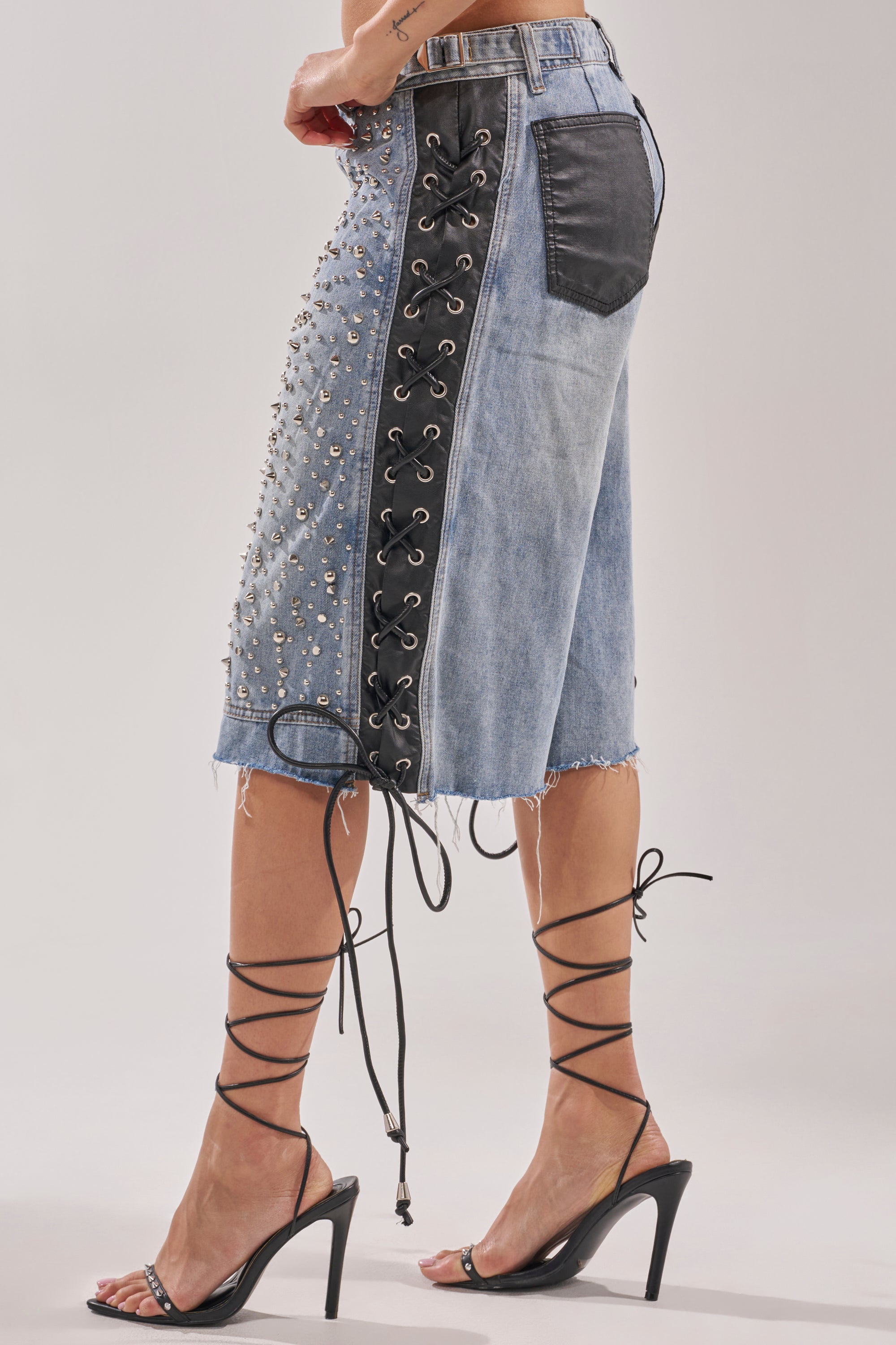 Wearing HONEY BABY BERMUDA SHORT, a high-waisted light-wash denim with black studs, leather pocket, lace-up front, and frayed hems, styled with strappy black heels. 4