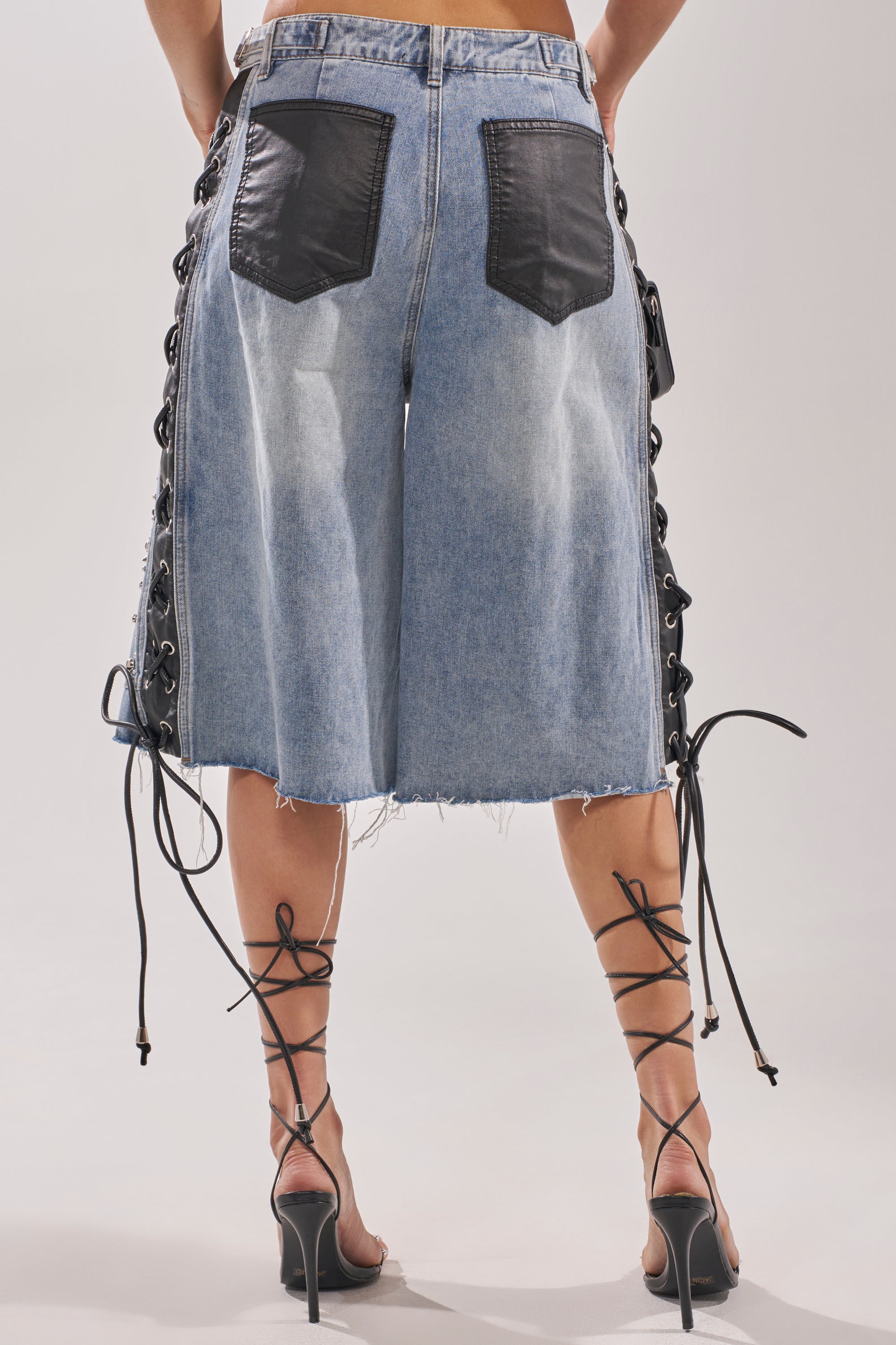 Wearing the HONEY BABY BERMUDA SHORT—knee-length, light blue lace-up denim with black leather back pockets and frayed hems—plus black strappy high-heeled sandals, this look serves a bold Y2K streetwear vibe from the back. 5