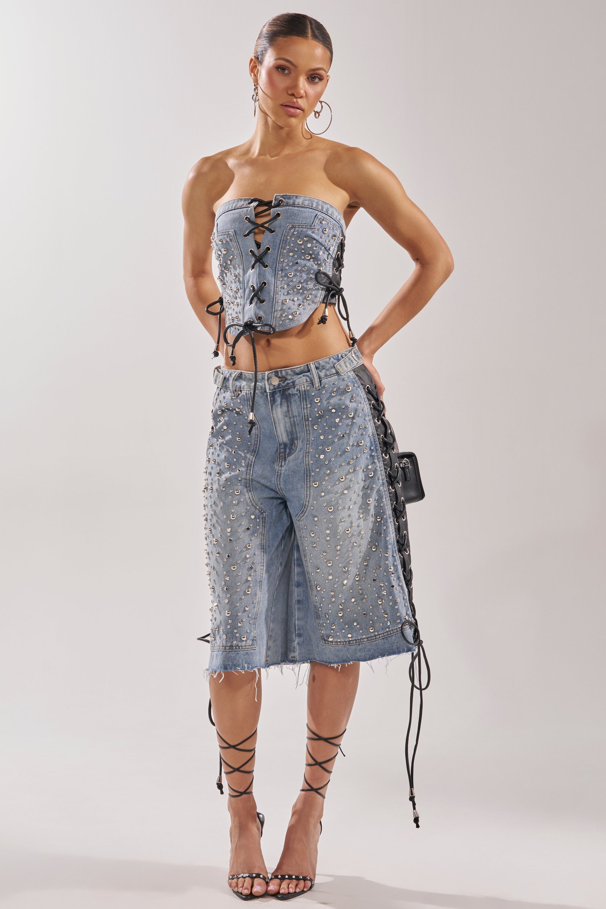 A woman wears the HONEY BABY BERMUDA SHORT with a denim strapless lace-up corset top, channeling Y2K streetwear. She completes her look with strappy black heels, hoop earrings, and a small black handbag. 6