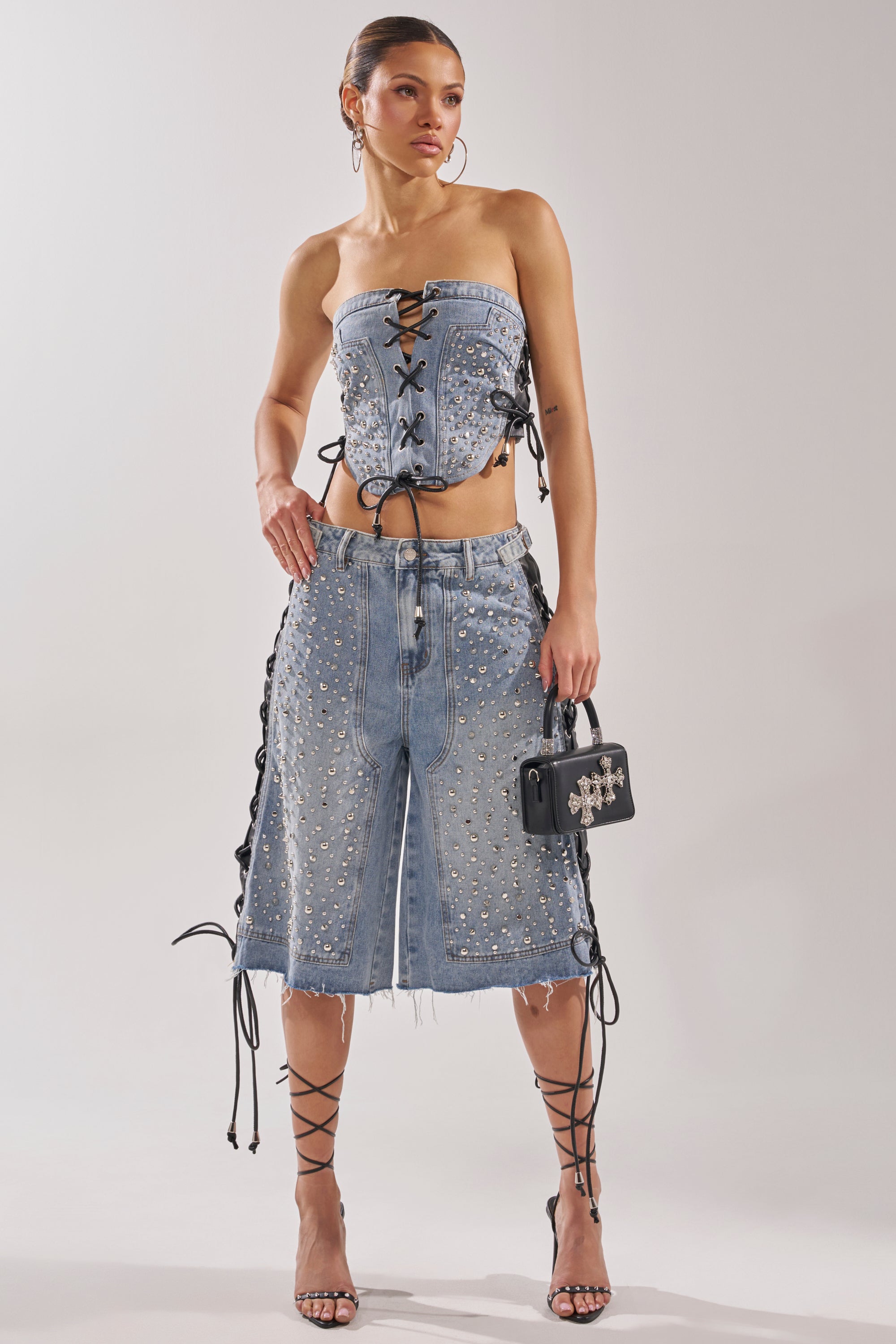 A woman models the HONEY BABY BERMUDA SHORT with a strapless denim top featuring lace-up sides, embracing Y2K streetwear. She finishes the outfit with strappy heels and a small black handbag. 7
