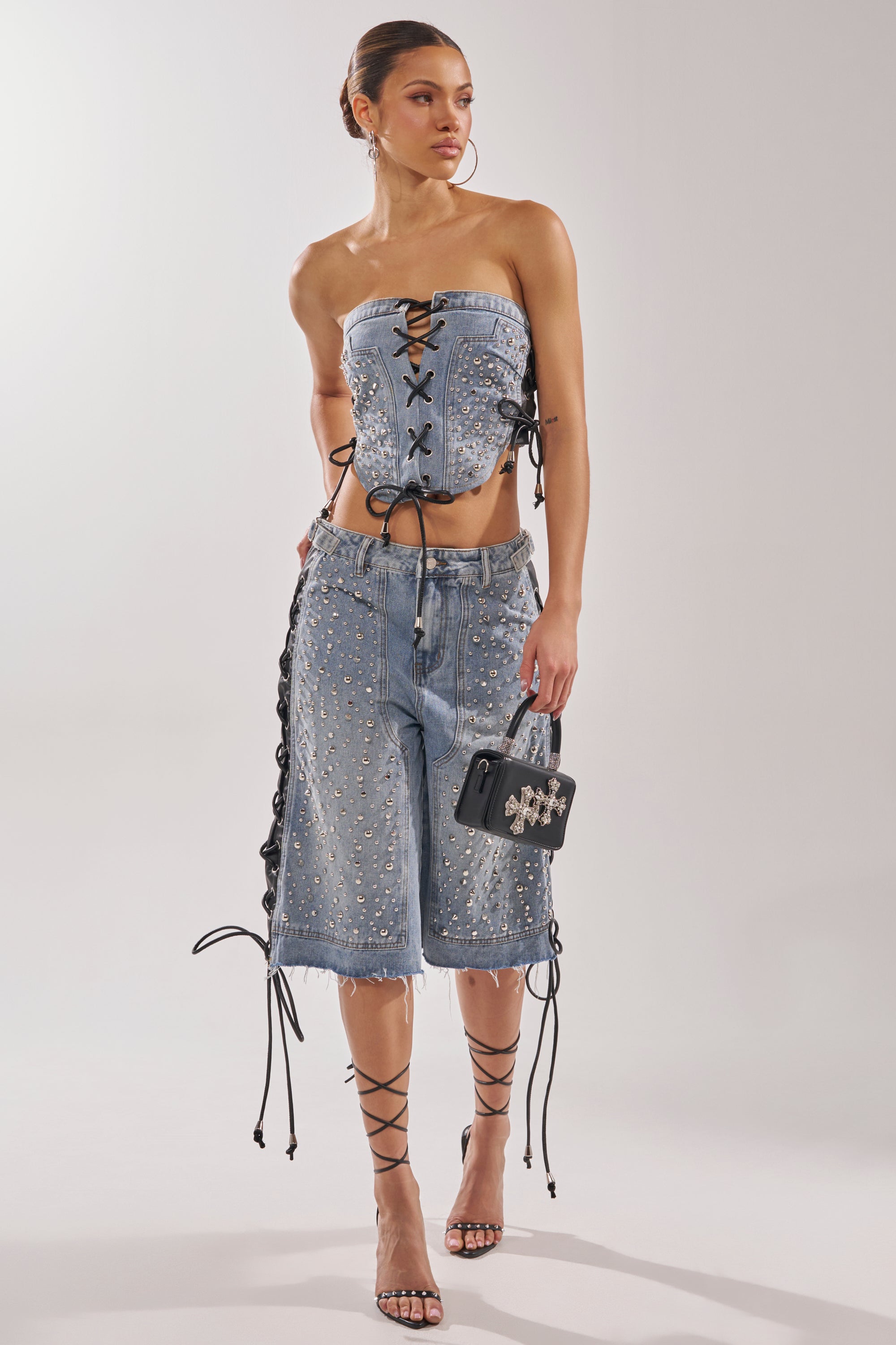 A woman wears a strapless lace-up denim corset, HONEY BABY BERMUDA SHORT inspired by Y2K streetwear, strappy heeled sandals, and carries a small black handbag. 8