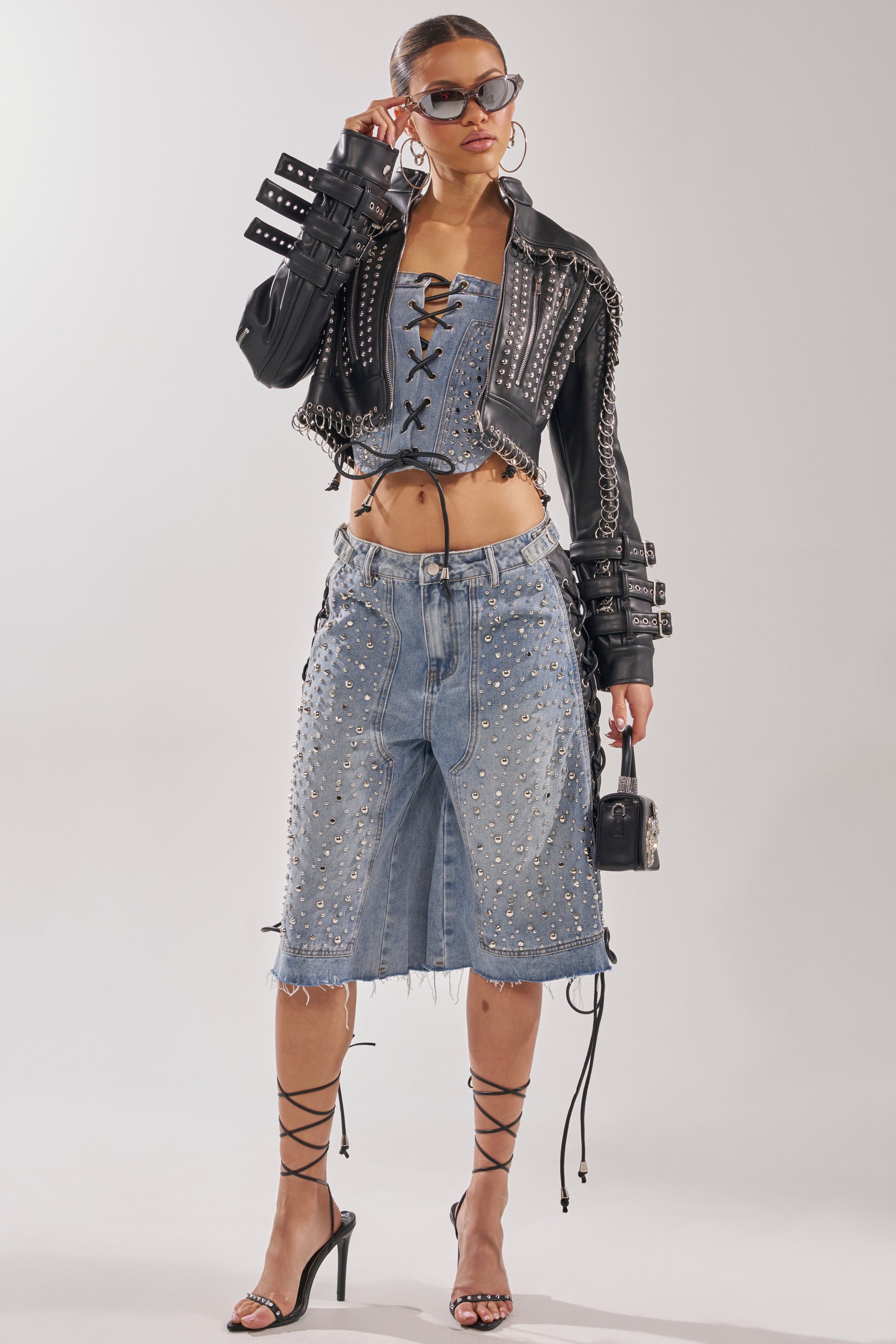 A stylish woman rocks the HONEY BABY LACE UP CORSET with a denim skirt, black buckle-detail leather jacket, strappy heels, sunglasses, hoop earrings, and a chic handbag—exuding fierce streetwear energy against a light backdrop. 9