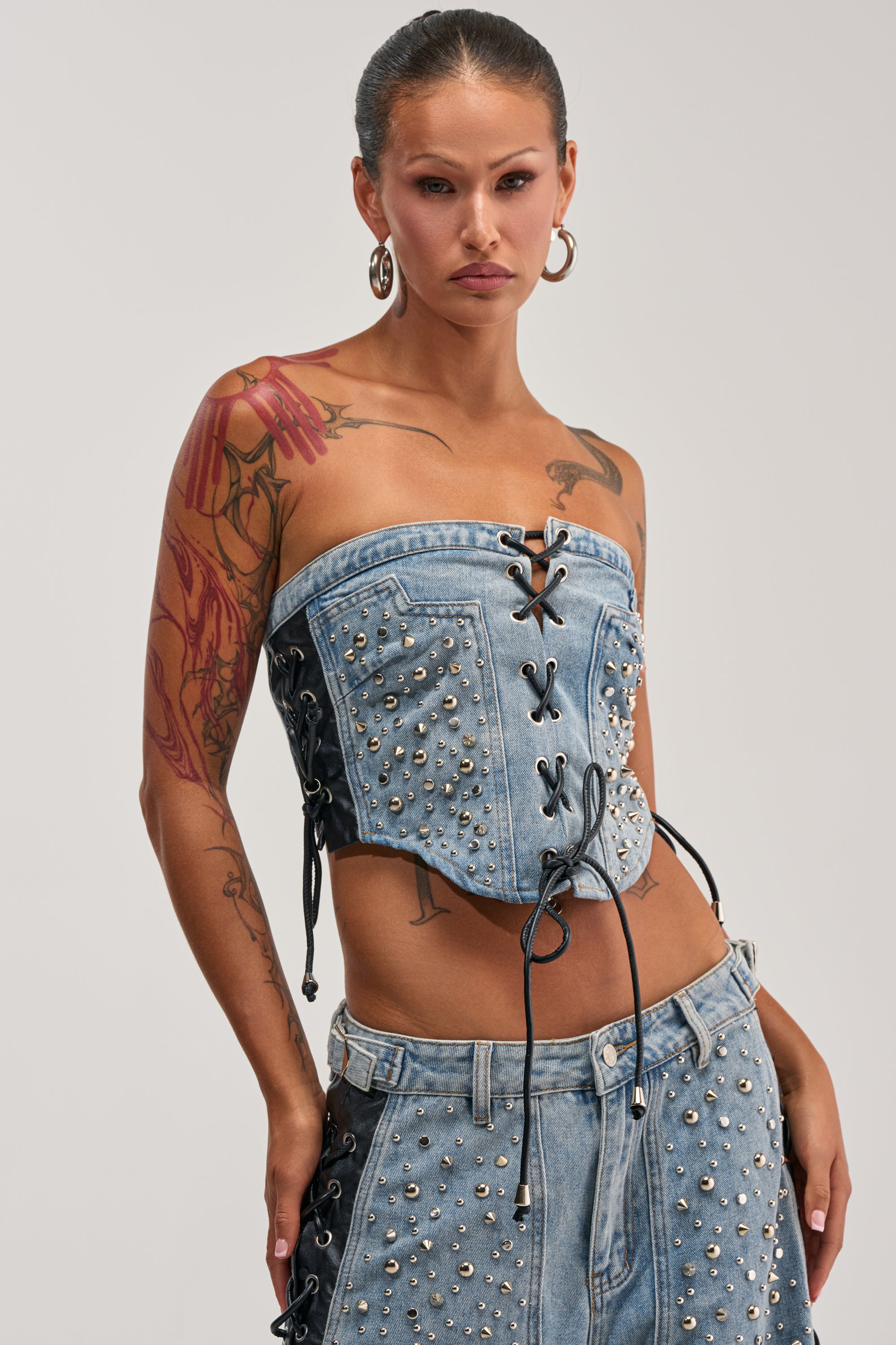 Wearing the HONEY BABY LACE UP CORSET—a strapless, lace-up denim top embellished with metal studs—paired with a matching denim skirt, this confident individual showcases their tattooed arms, hoop earrings, and slicked-back hair.