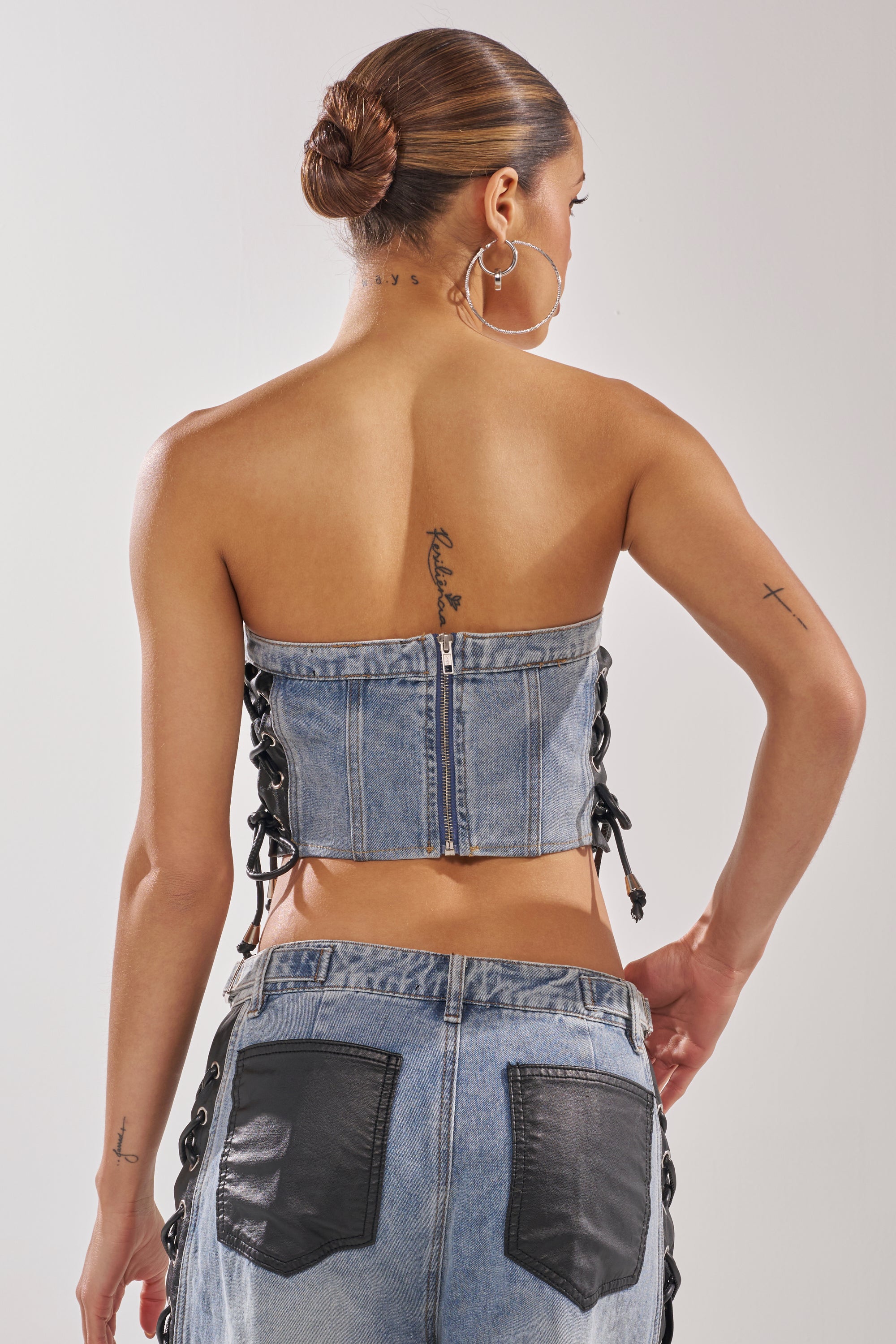 A woman with tattoos and hoop earrings faces away from the camera, wearing the HONEY BABY LACE UP CORSET and matching jeans featuring black side lacing and pocket patches, her hair in a sleek bun for bold streetwear style. 4