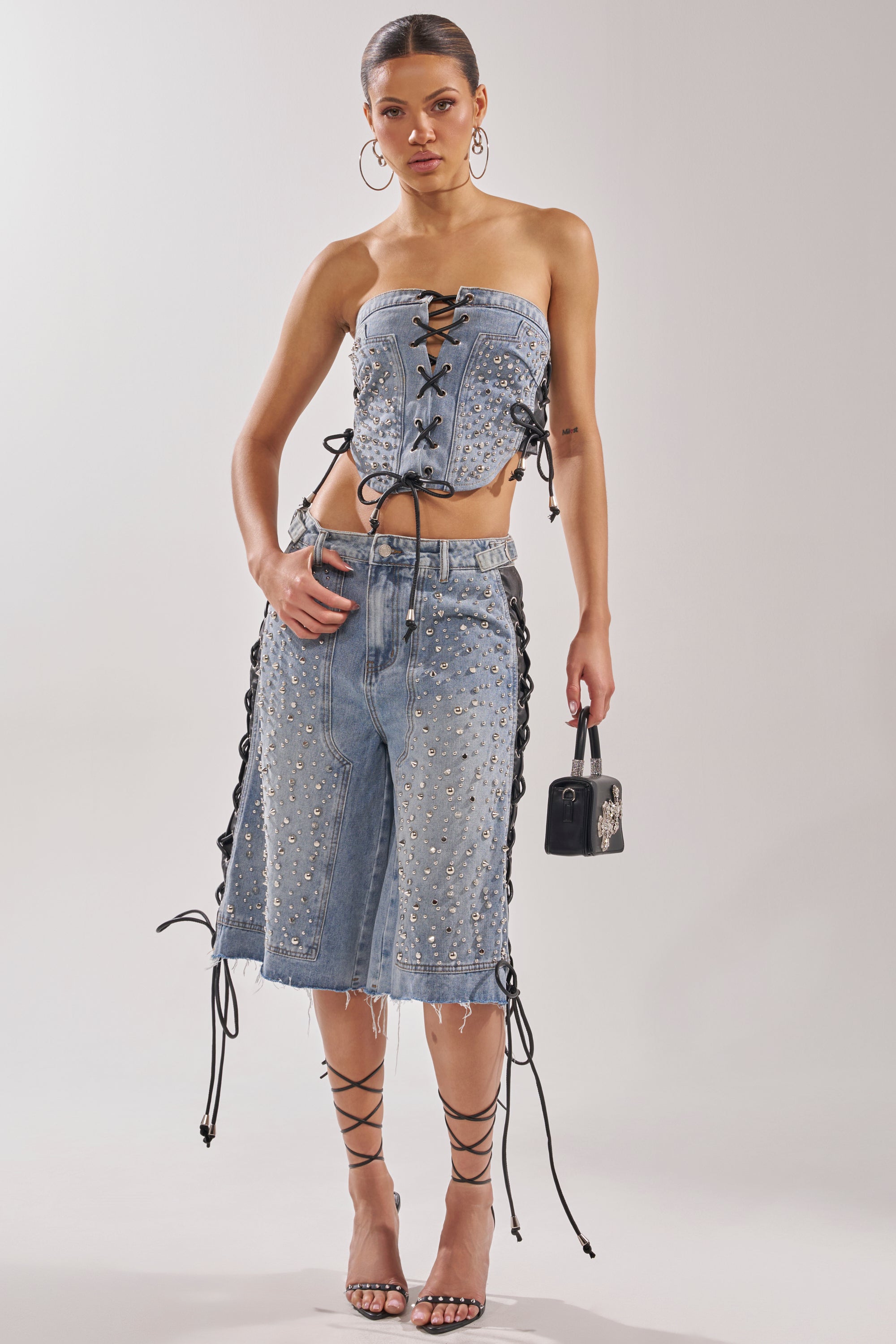A woman wears the HONEY BABY LACE UP CORSET with matching studded lace-up shorts, strappy heels, hoop earrings, and carries a small black handbag. 5