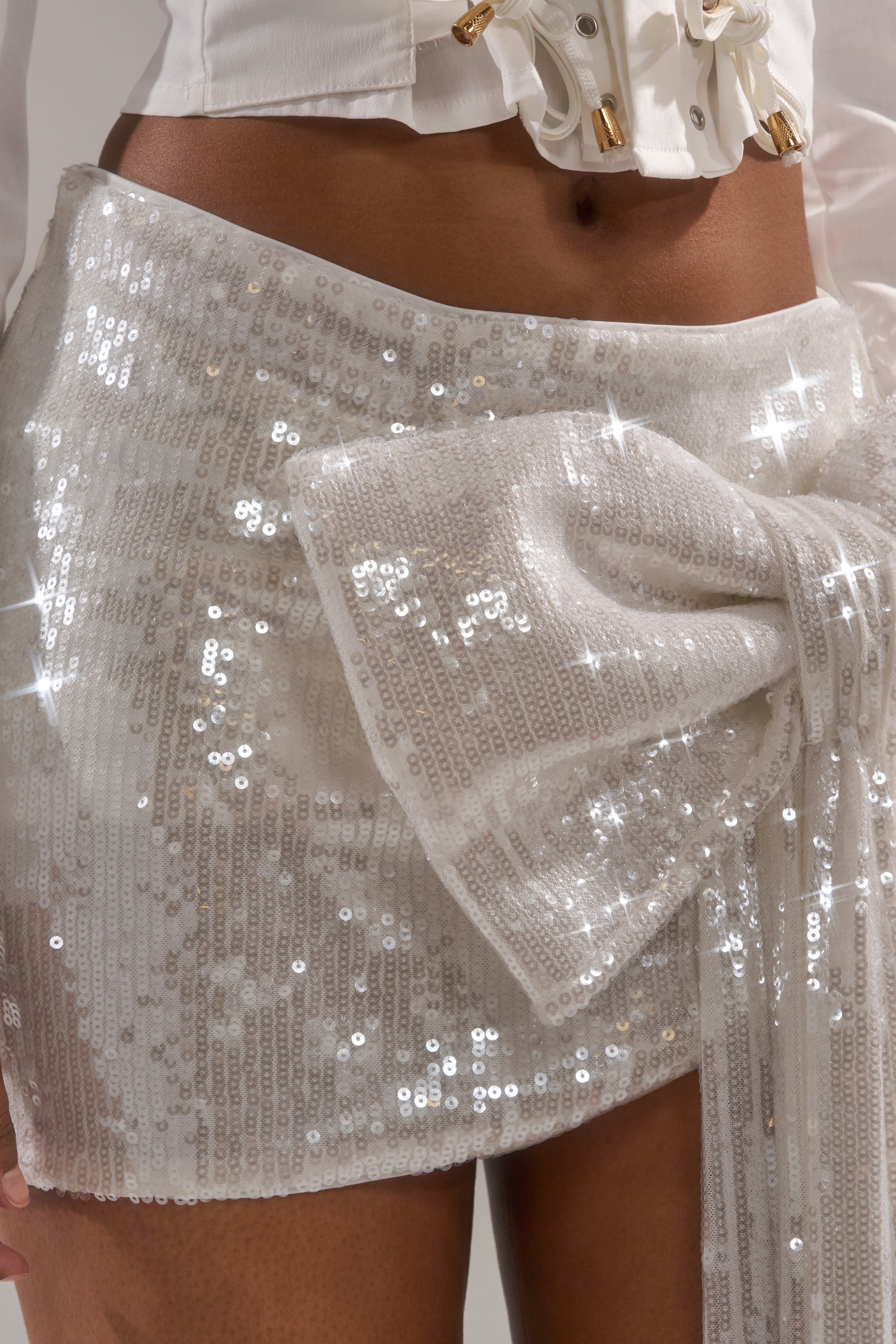 A close-up of someone wearing the SHINING DOWN ON YOU SEQUIN MINI SKIRT, featuring white sequins and a sparkling side bow—ideal for holiday parties. This high-rise mini skirt shines under the light and is styled with a white corset top.