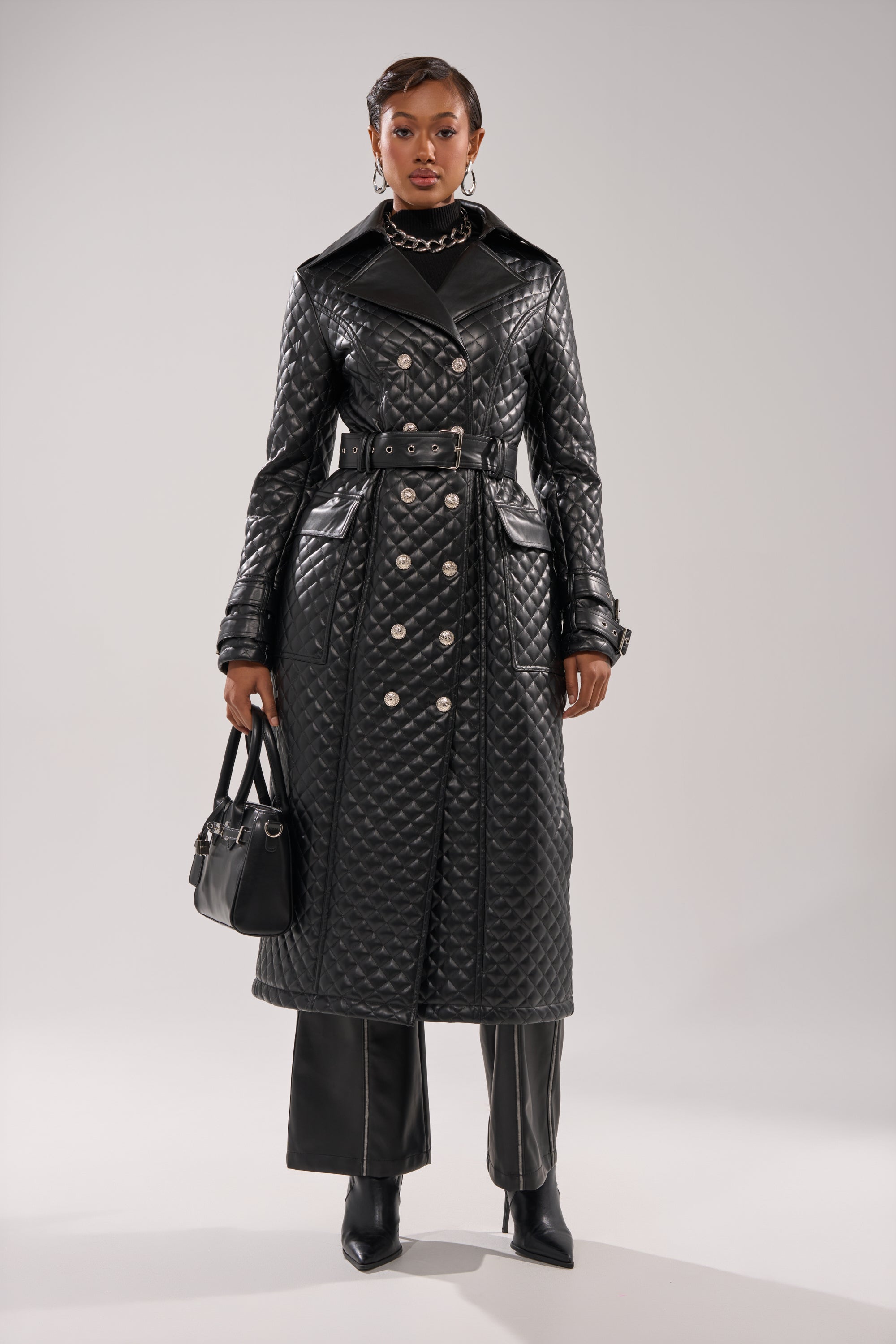 A woman stands against a plain background wearing the MADISON AVE QUILTED TRENCH, styled with black wide-leg pants, pointed boots, a black handbag, earrings, and layered necklaces.