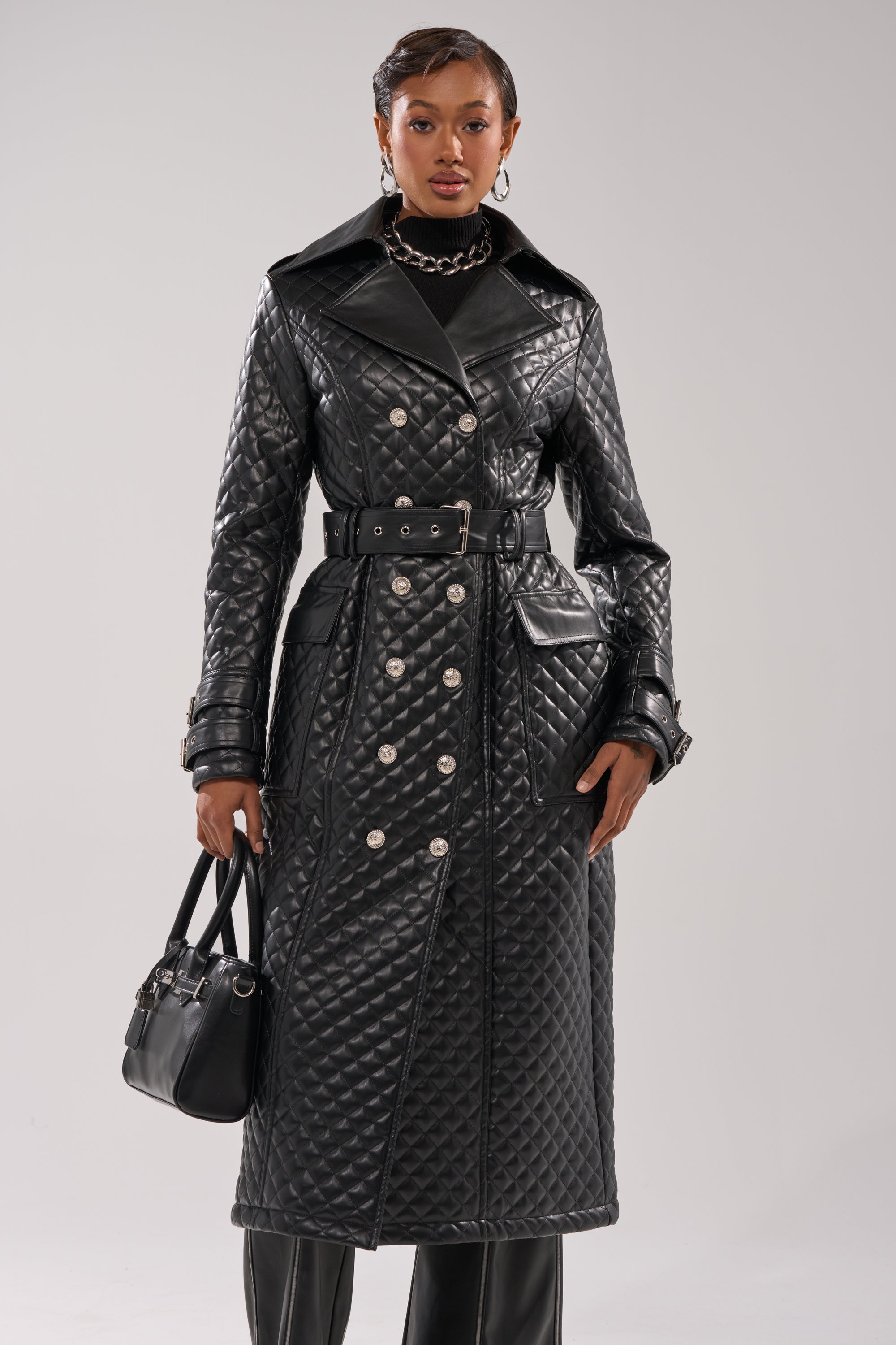 A person with short hair wears the MADISON AVE QUILTED TRENCH—a long, black belted coat with silver buttons—styled with a black handbag, statement earrings, and a chunky necklace against a plain light gray background.