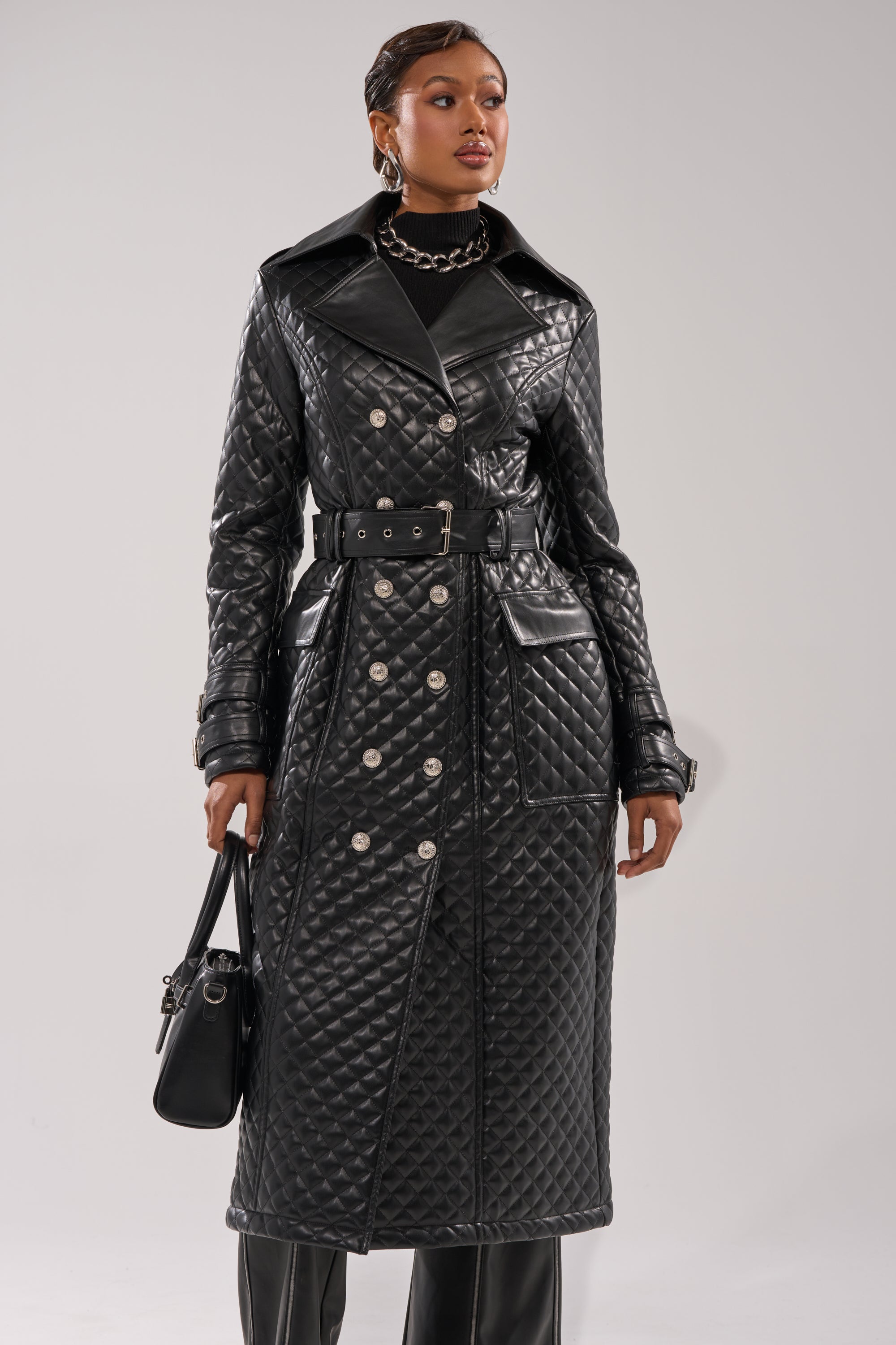 Wearing the MADISON AVE QUILTED TRENCH, a woman stands against a plain background. The long black quilted coat features a belt and silver buttons, paired with a black handbag and chunky silver jewelry for a stylish look.