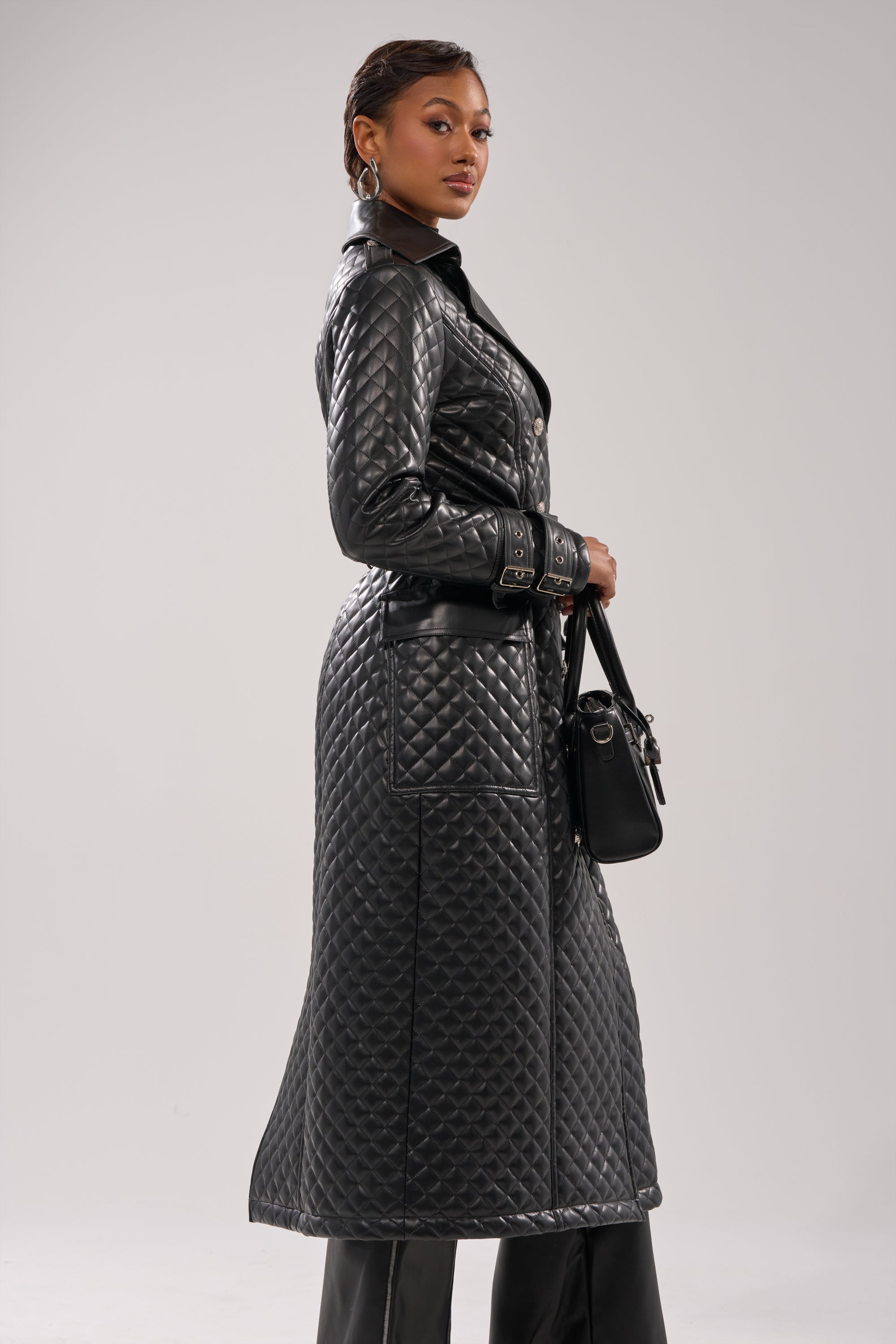 A woman models the MADISON AVE QUILTED TRENCH, a long trench coat with a belted waist, paired with a black handbag and hoop earrings against a plain, light background.