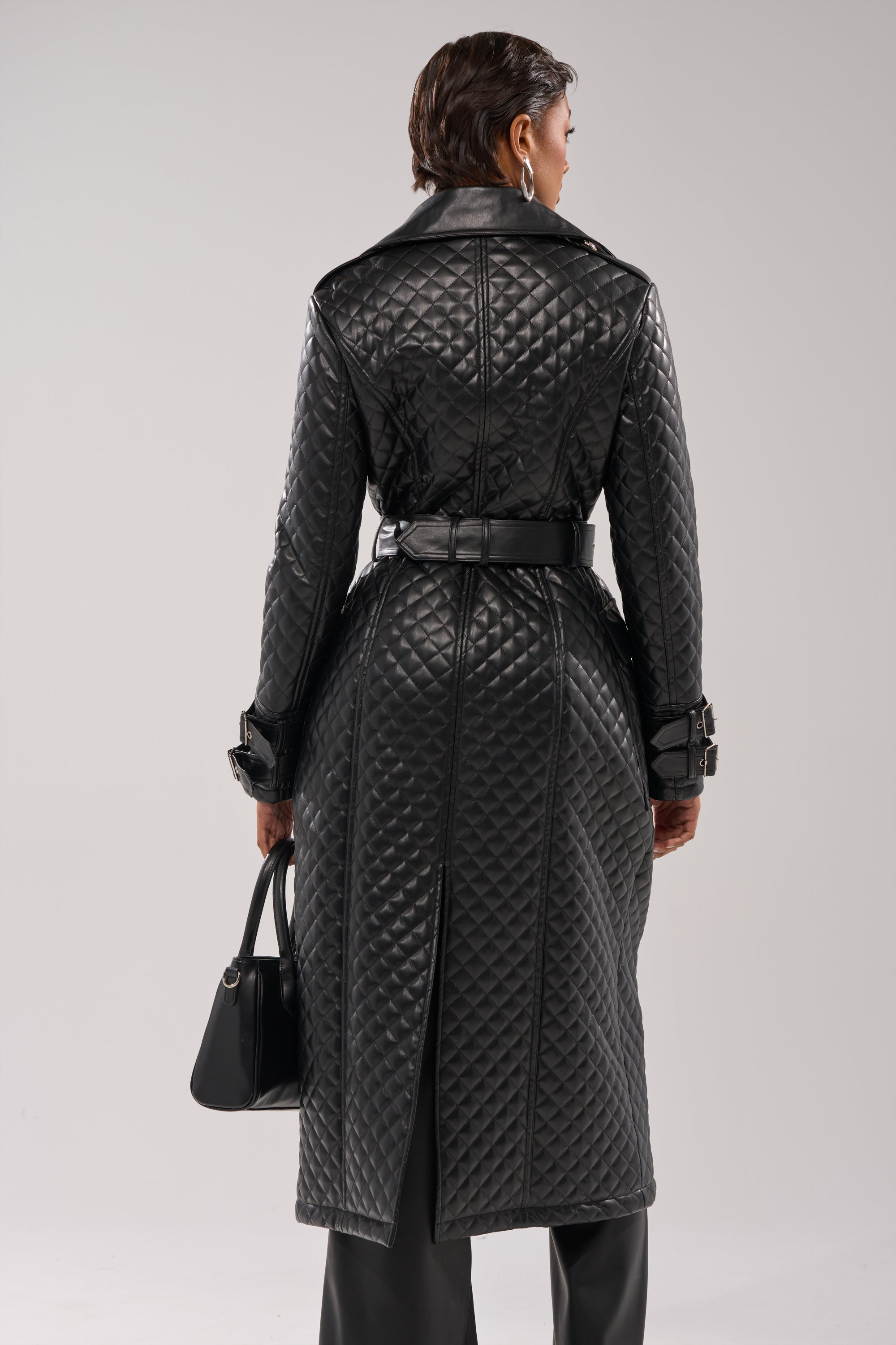 A person with neat, short dark hair holds a black handbag and wears the MADISON AVE QUILTED TRENCH—a long black belted coat with buckled cuffs. They face away against a plain, light background.