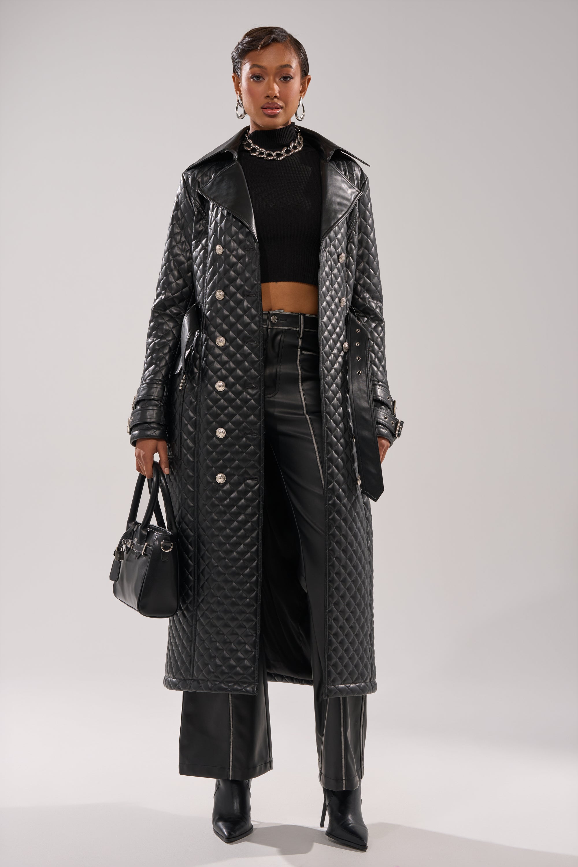 A woman stands confidently in the MADISON AVE QUILTED TRENCH over a black crop top and leather pants, accessorizing with a black handbag, boots, and a chunky chain necklace.