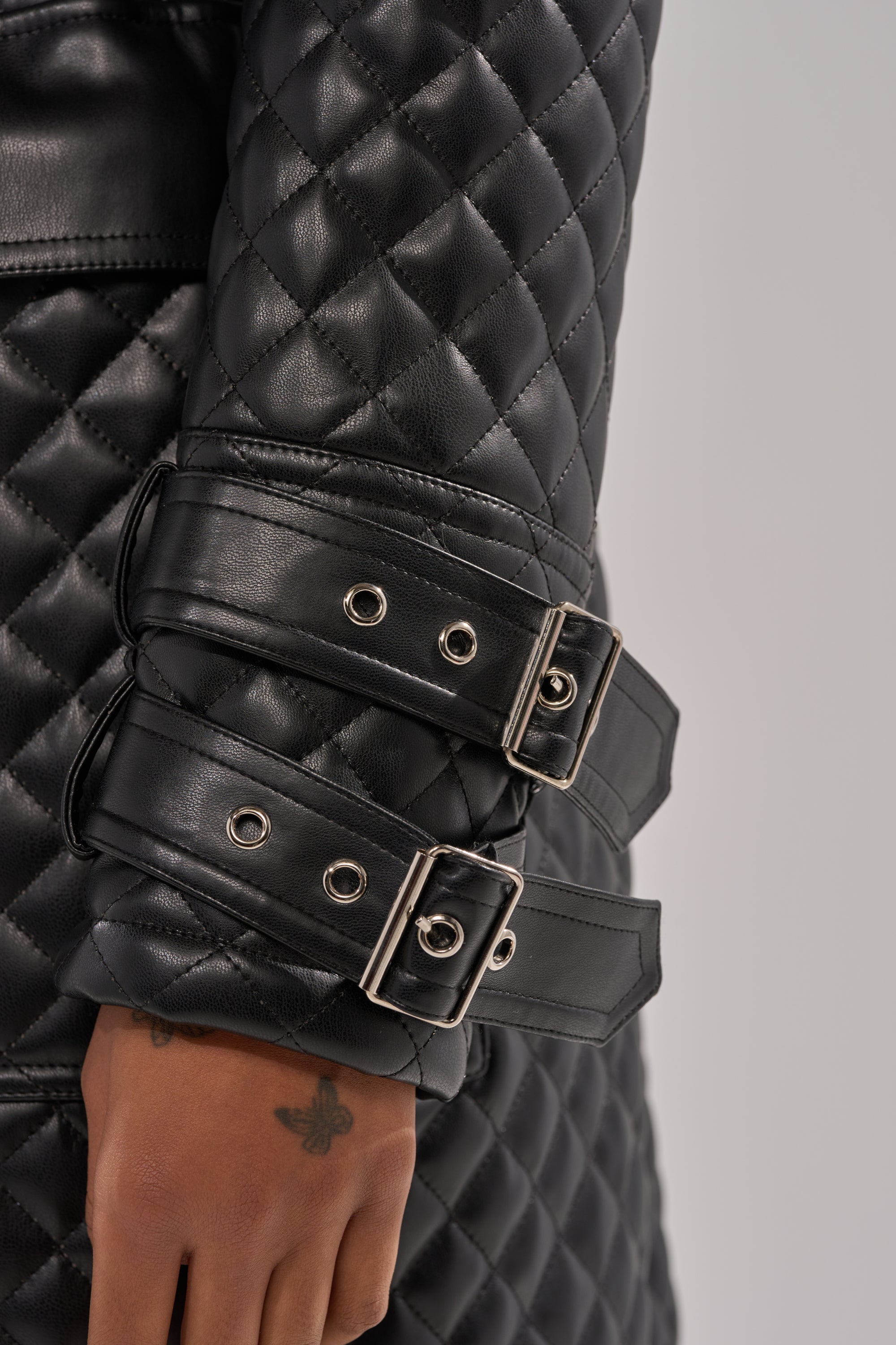 Close-up of a person's arm in the MADISON AVE QUILTED TRENCH, featuring black quilted fabric and two silver-buckled straps on the sleeve. A small bee tattoo is visible on the person's hand.