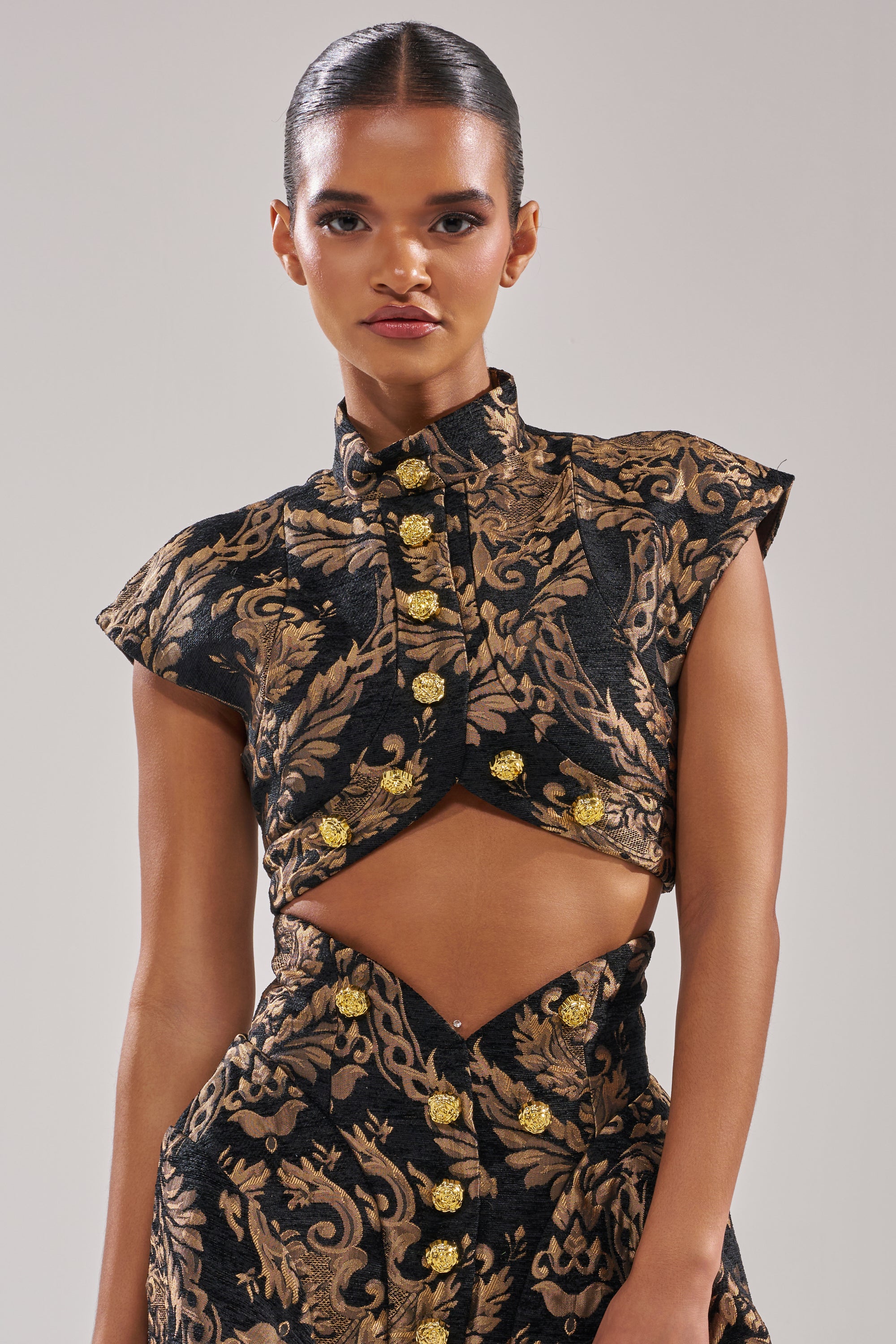 A woman wears the KAYLA BROCADE LONG SLEEVE BLOUSE in black and gold brocade with gold buttons and an asymmetrical midriff cutout, creating a chic holiday party look against a plain light background.