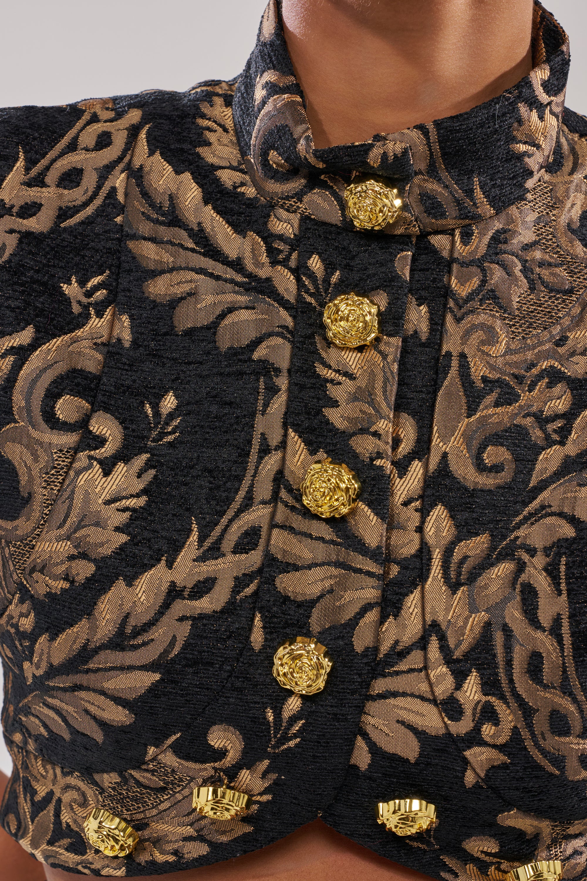 A close-up of a person wearing the KAYLA BROCADE LONG SLEEVE BLOUSE, a black and gold top featuring ornate floral patterns and shiny gold buttons—an elegant, festive choice for holiday parties.