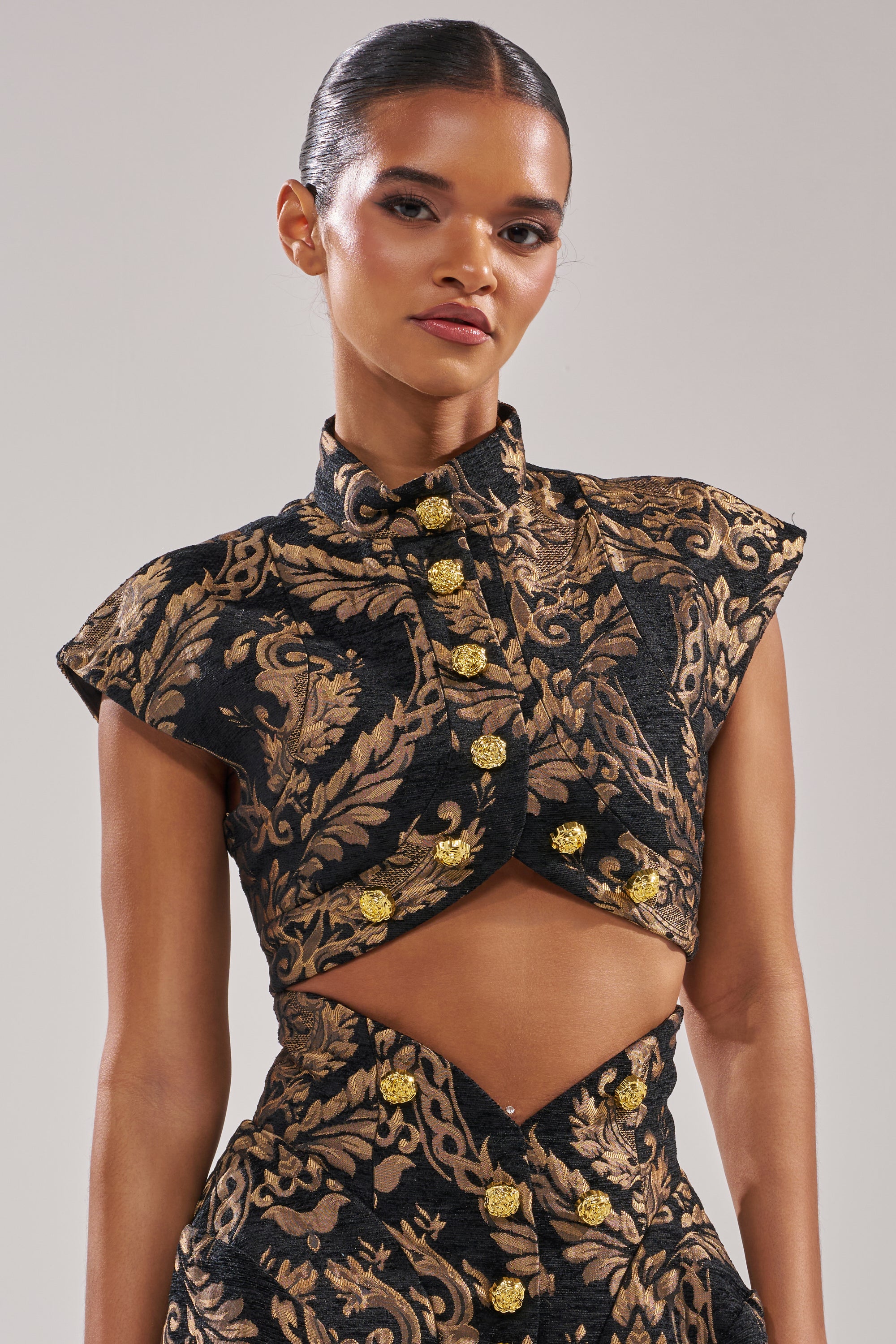 A woman models the KAYLA BROCADE LONG SLEEVE BLOUSE, featuring black and gold ornate patterns, gold buttons, cap sleeves, and a waist cutout. Her slicked-back hair and natural makeup create a chic look—ideal for holiday parties.