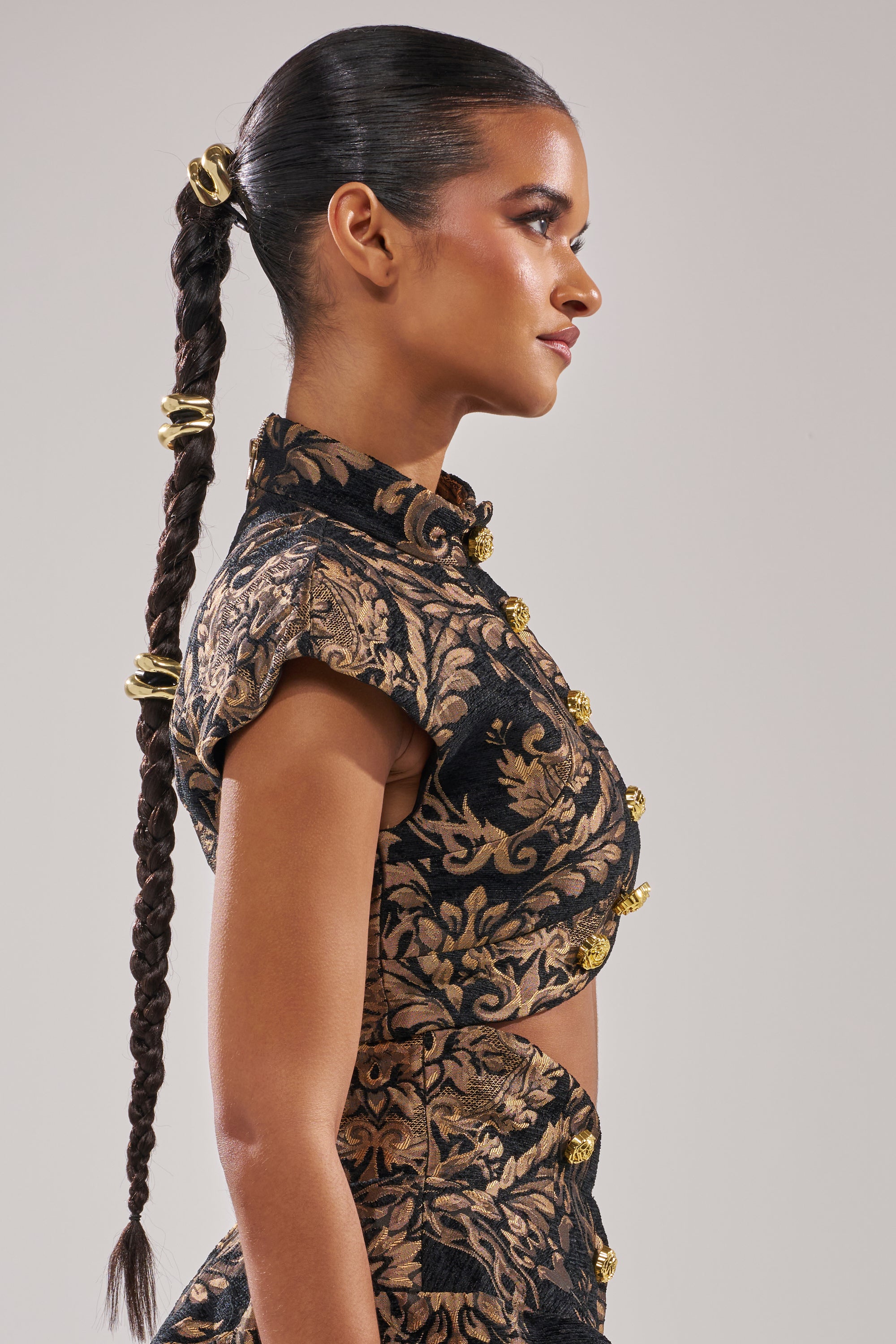 A woman with a sleek, braided ponytail and gold bands is shown in profile wearing the KAYLA BROCADE LONG SLEEVE BLOUSE—black with metallic gold accents—an elegant holiday party option, posed against a plain background.