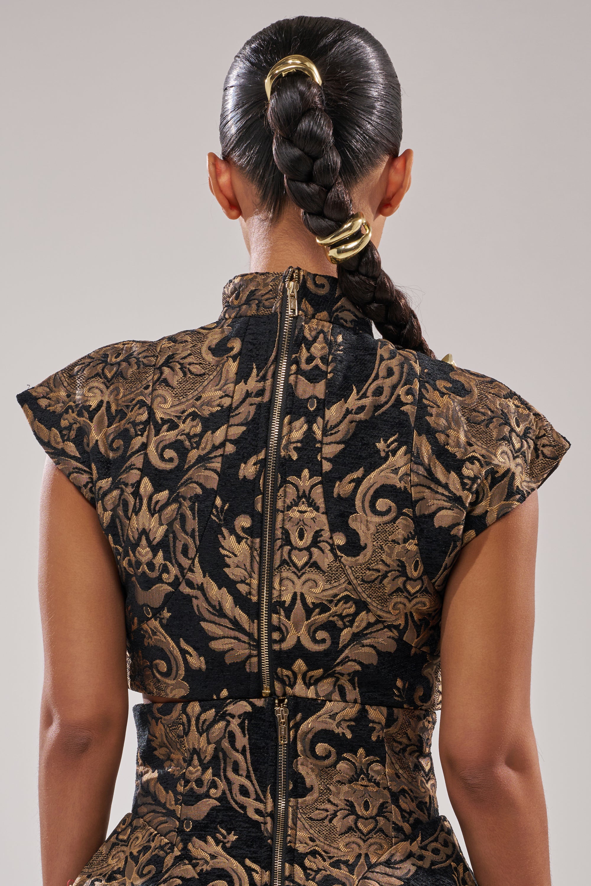 A person seen from behind wears a jacquard high-collared dress with ornate gold and black florals. Their braids, secured with gold bands, evoke the style of the AKIRA Label KAYLA BROCADE LONG SLEEVE BLOUSE.