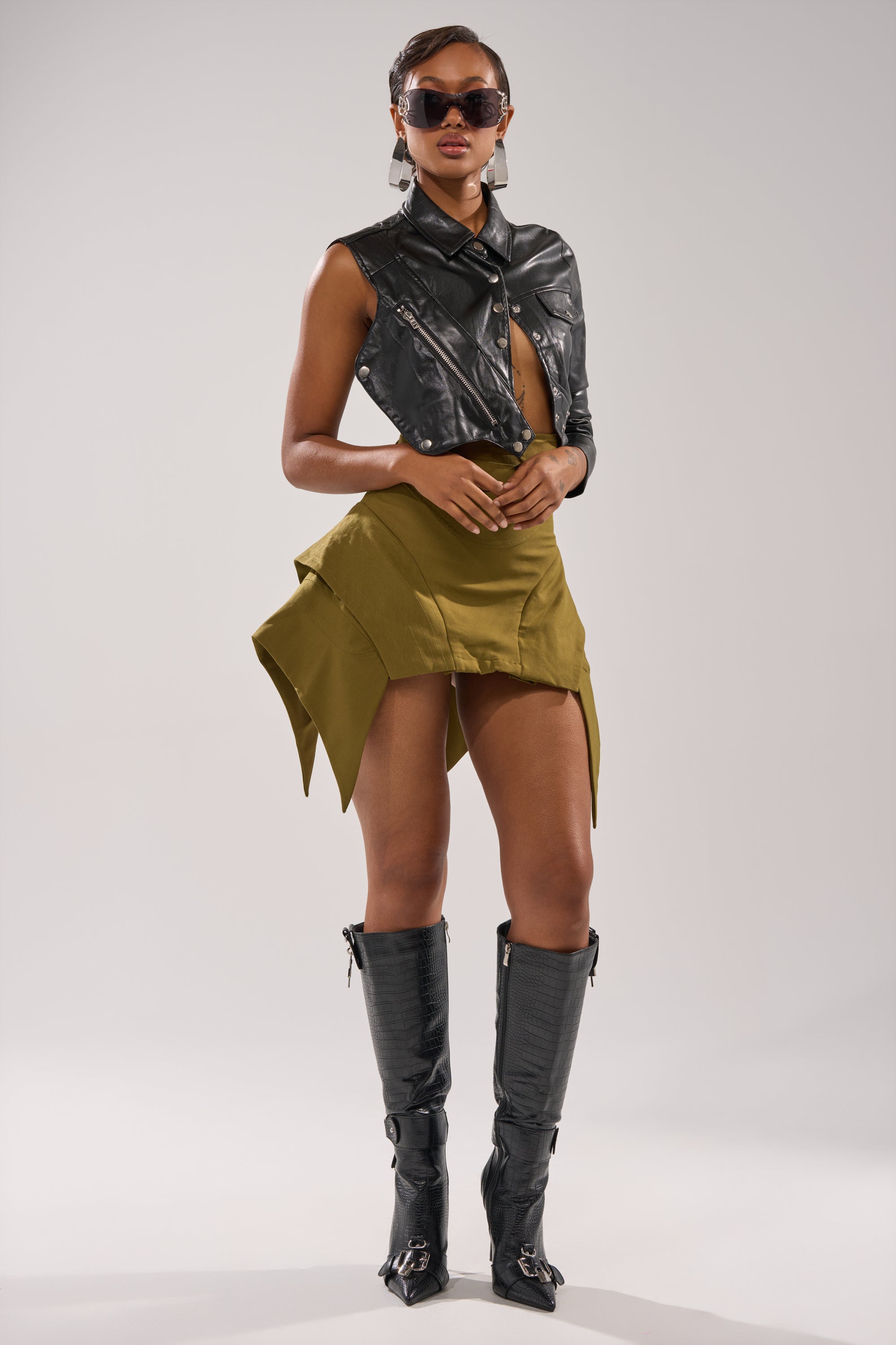 A woman poses confidently in a sleeveless black leather jacket, tall black boots, hoop earrings, and dark sunglasses, styled with the DONT LOOK BACK MINI SKIRT IN OLIVE against a plain light background. 5