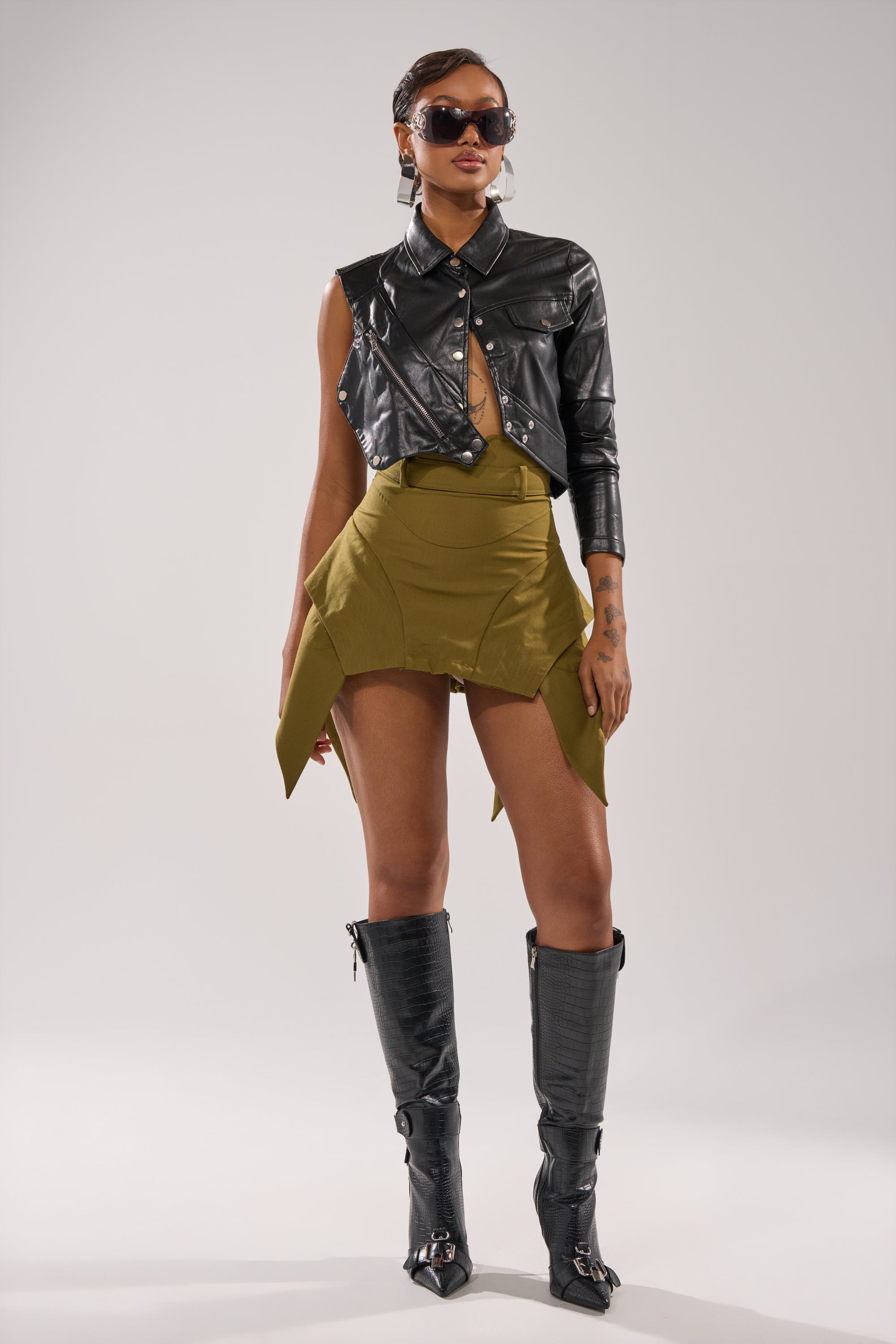 A woman stands confidently against a plain light background, wearing the DONT LOOK BACK MINI SKIRT IN OLIVE styled with a black cropped leather jacket, knee-high boots, and sunglasses. 7