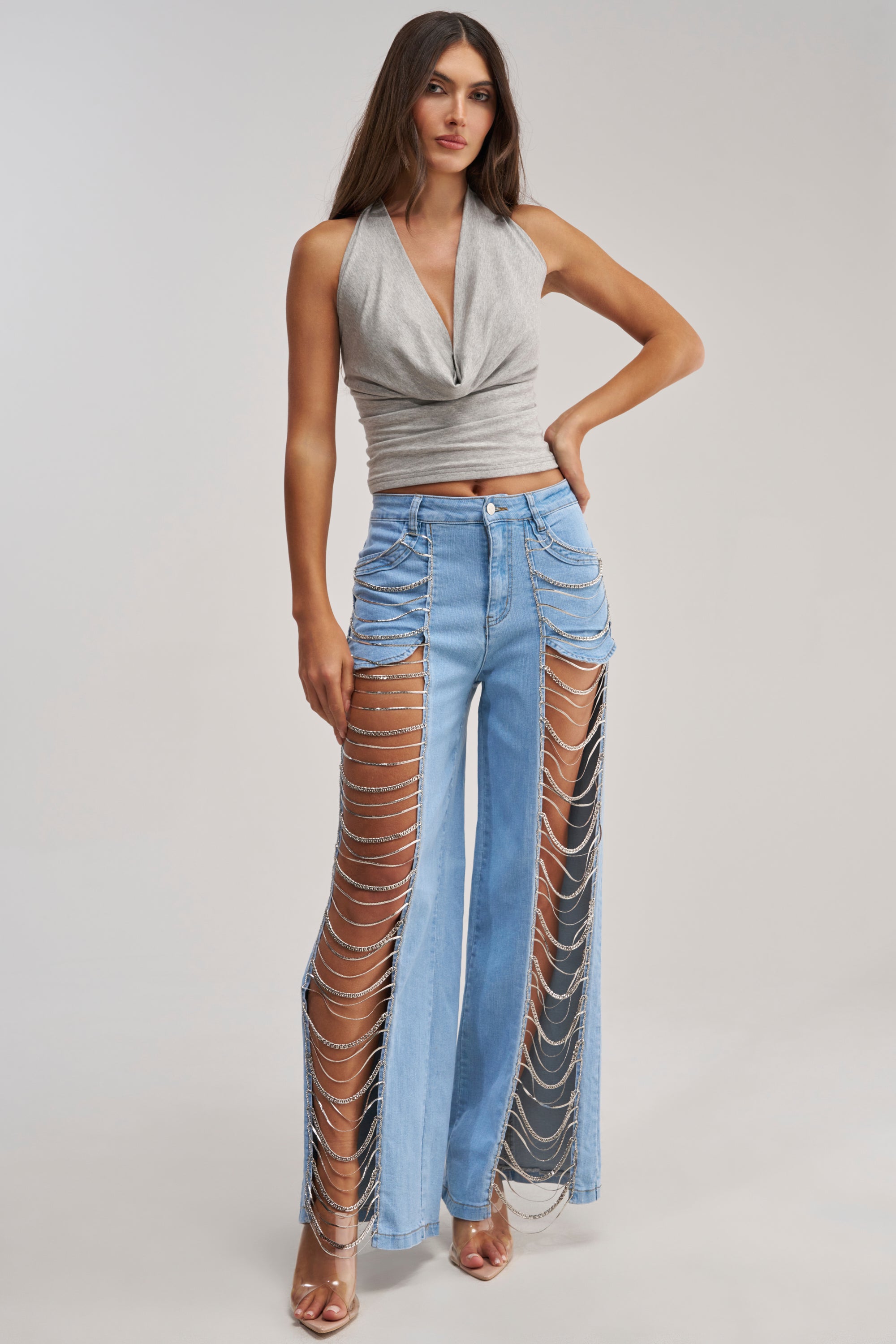 A woman wears CHAINS FOR DAYS RELAXED DENIM—light blue cutout jeans with draped silver chains—and a sleeveless gray halter top, paired with clear high-heeled sandals. She has long brown hair.