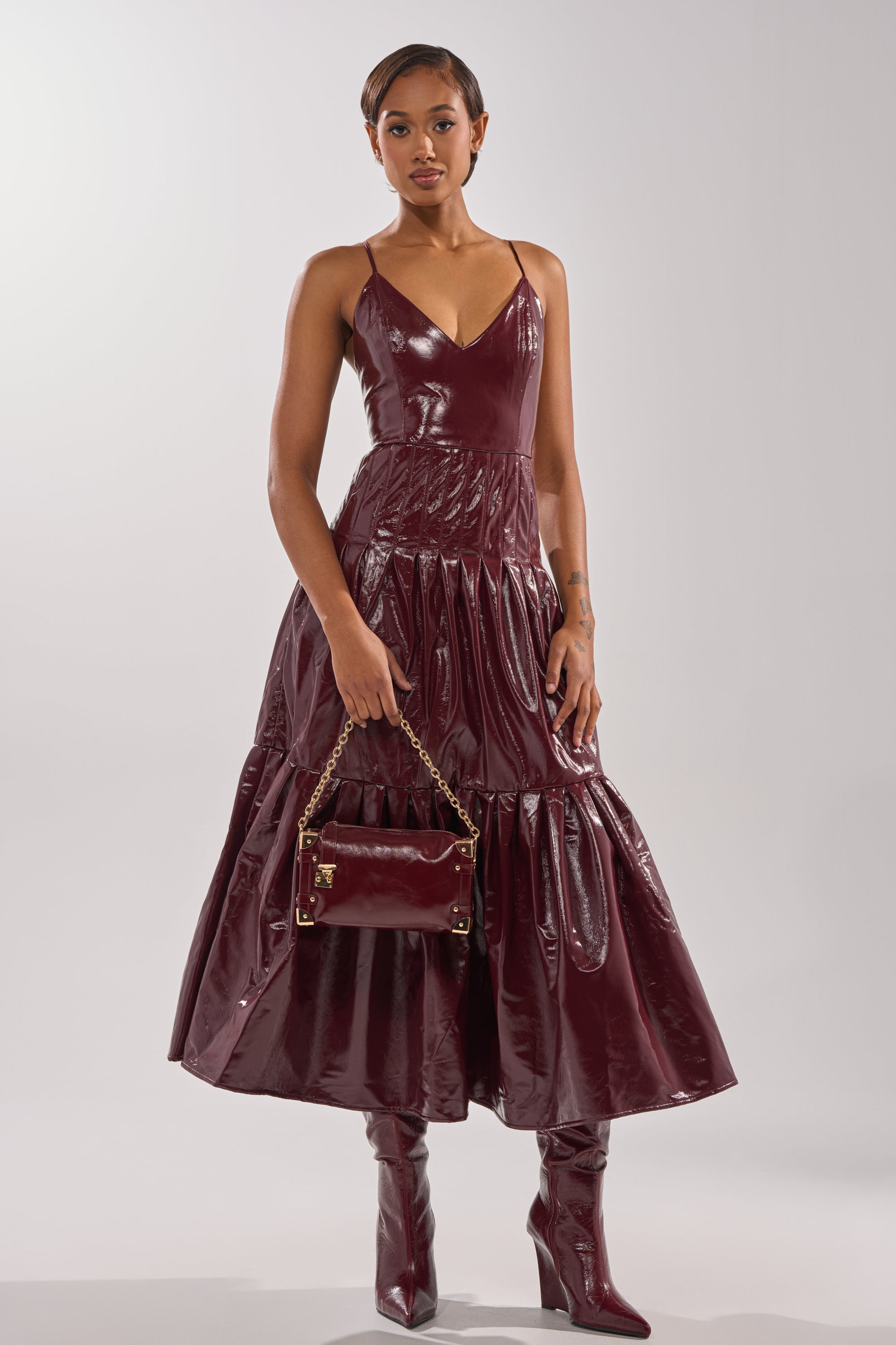 A woman wearing the RACHEL PU MAXI DRESS in shiny burgundy faux leather, paired with matching knee-high heeled boots and a small burgundy gold-chain handbag, stands against a plain light background.