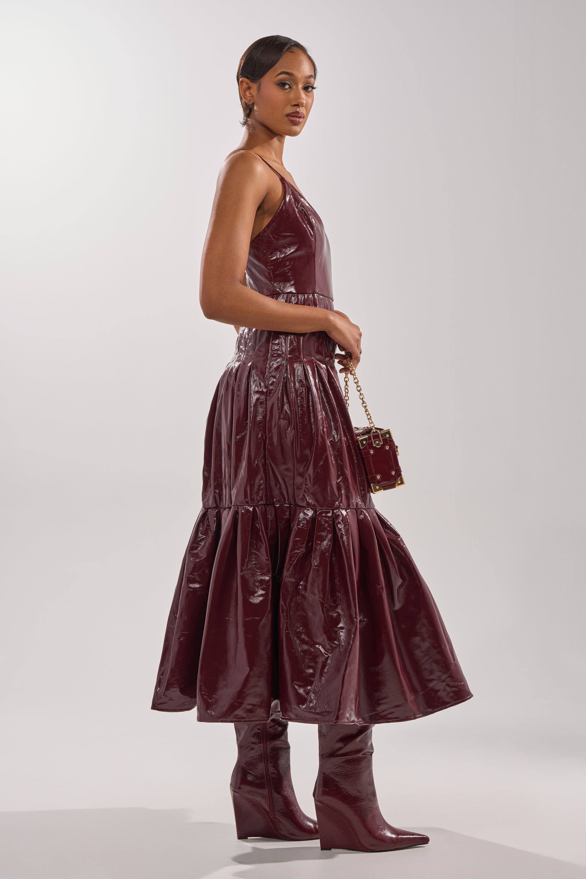 A woman in profile wears the RACHEL PU MAXI DRESS, a sleeveless, shiny burgundy faux leather dress with a tiered skirt, matching knee-high boots, and holds a small square handbag against a plain light background.