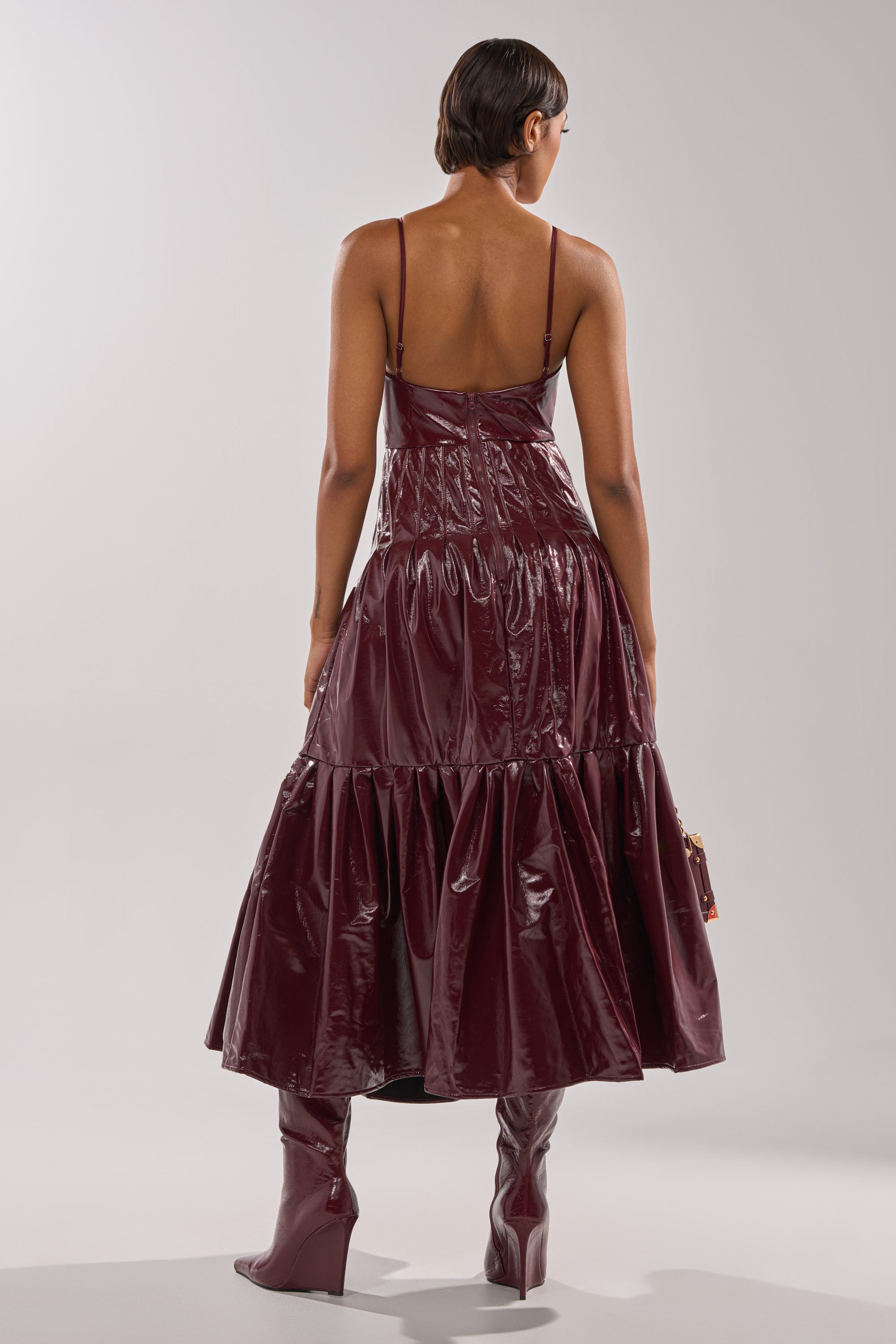 A person with short hair is shown from behind wearing the RACHEL PU MAXI DRESS, a shiny deep burgundy faux leather dress with thin straps and a tiered skirt, paired with matching high boots.