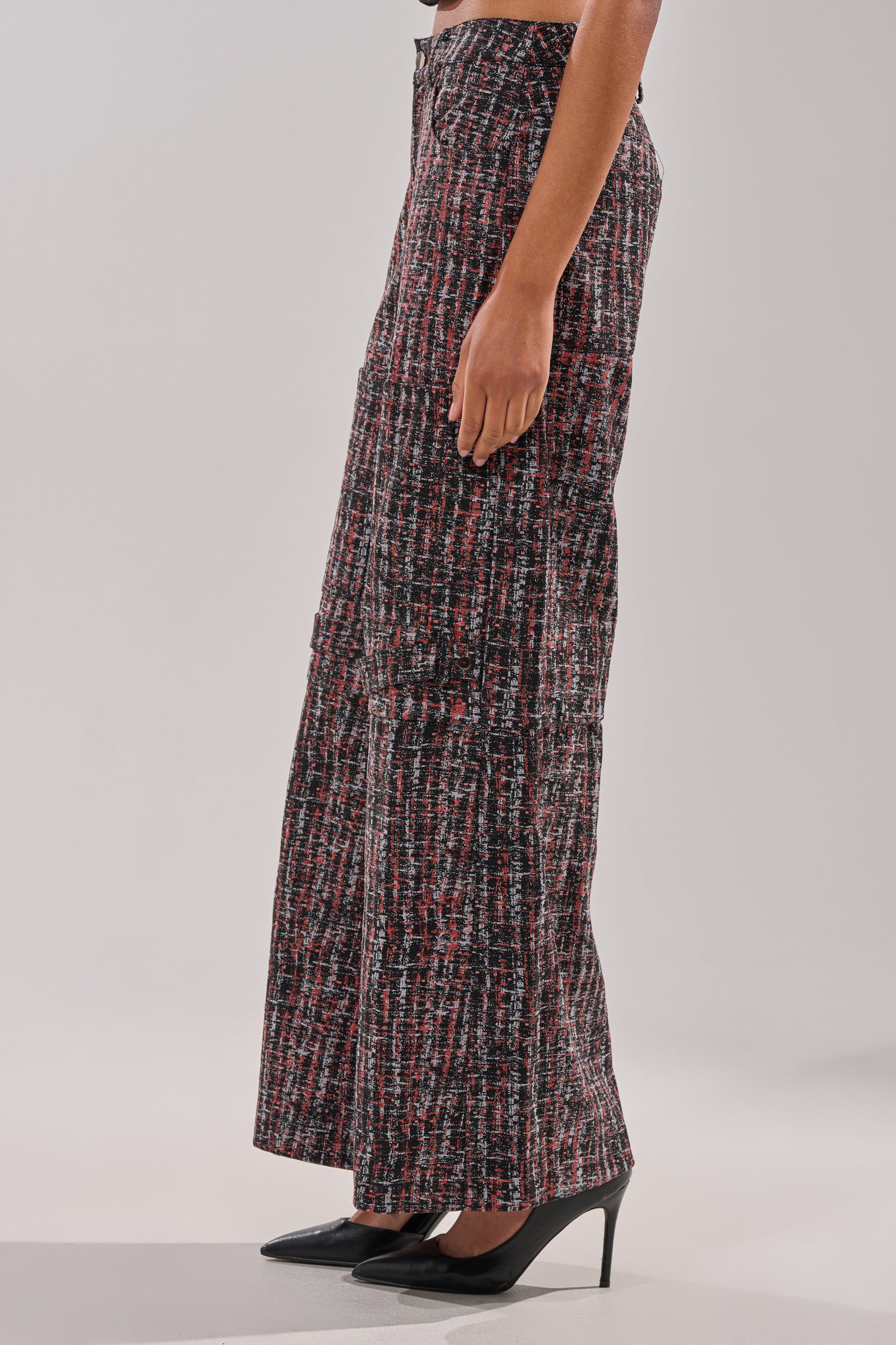 A person stands in profile wearing the WORK IT FAUX LEATHER PLAID TROUSER in black, red, and white plaid, styled with black pointed-toe heels against a plain light gray background. 3