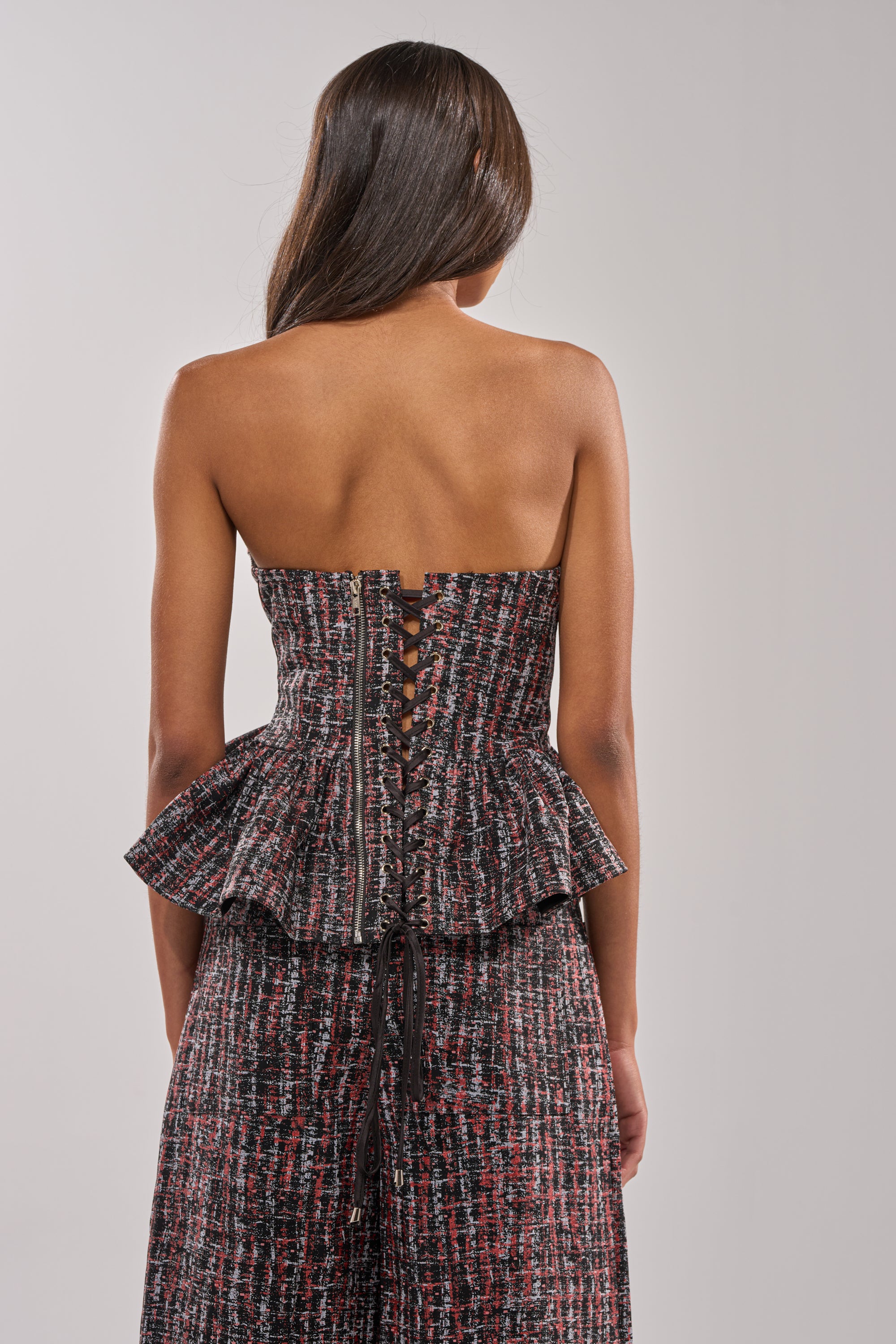 A woman stands with her back to the camera, wearing the WORK IT FAUX LEATHER PLAID CORSET featuring a strapless design, corseted back with black ties, textured plaid fabric, and a peplum flare for a chic silhouette. 3