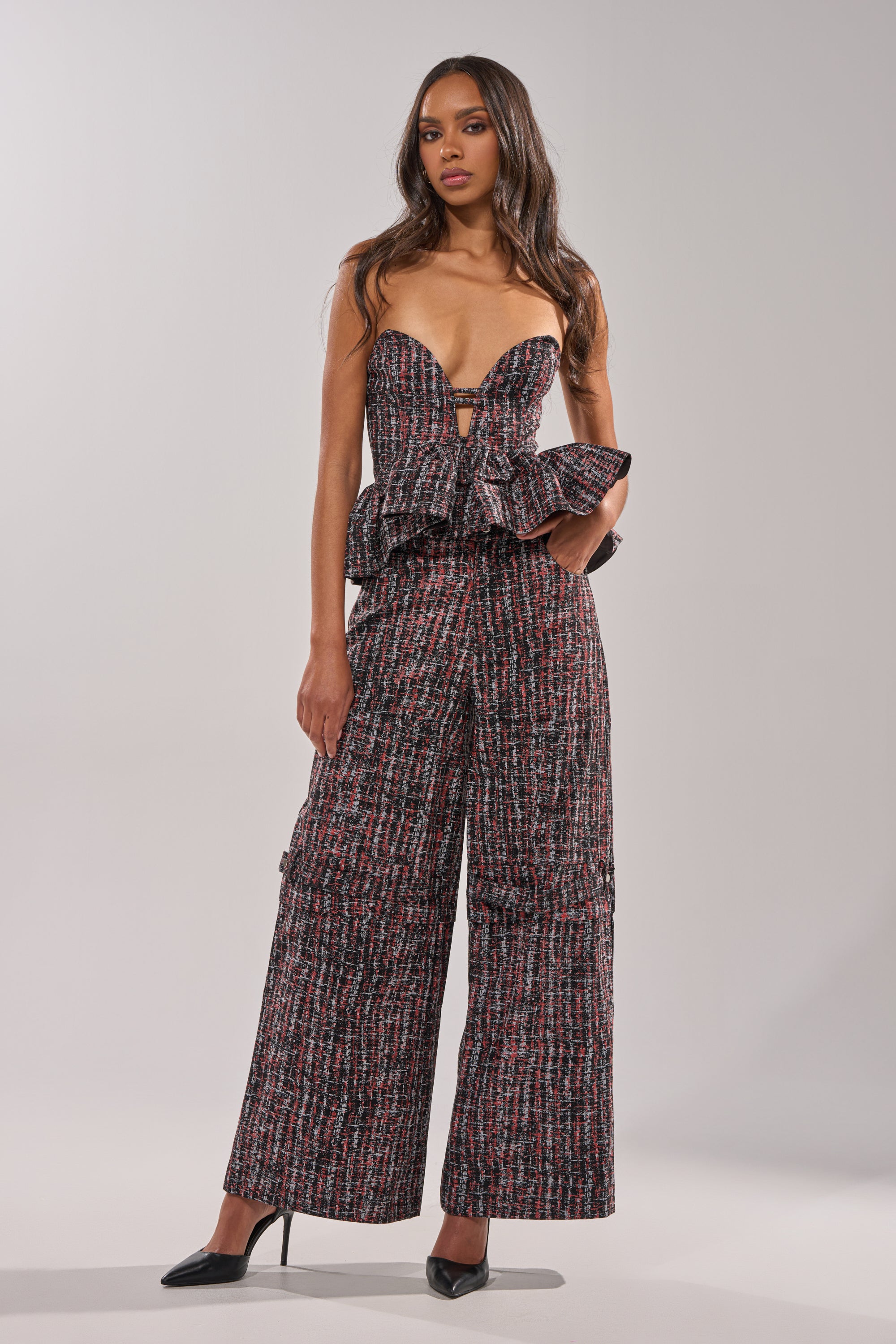 A woman wears the WORK IT FAUX LEATHER PLAID CORSET with a structured sweetheart neckline and matching wide-leg pants in red, black, and white plaid, paired with black heels against a simple light backdrop. 4