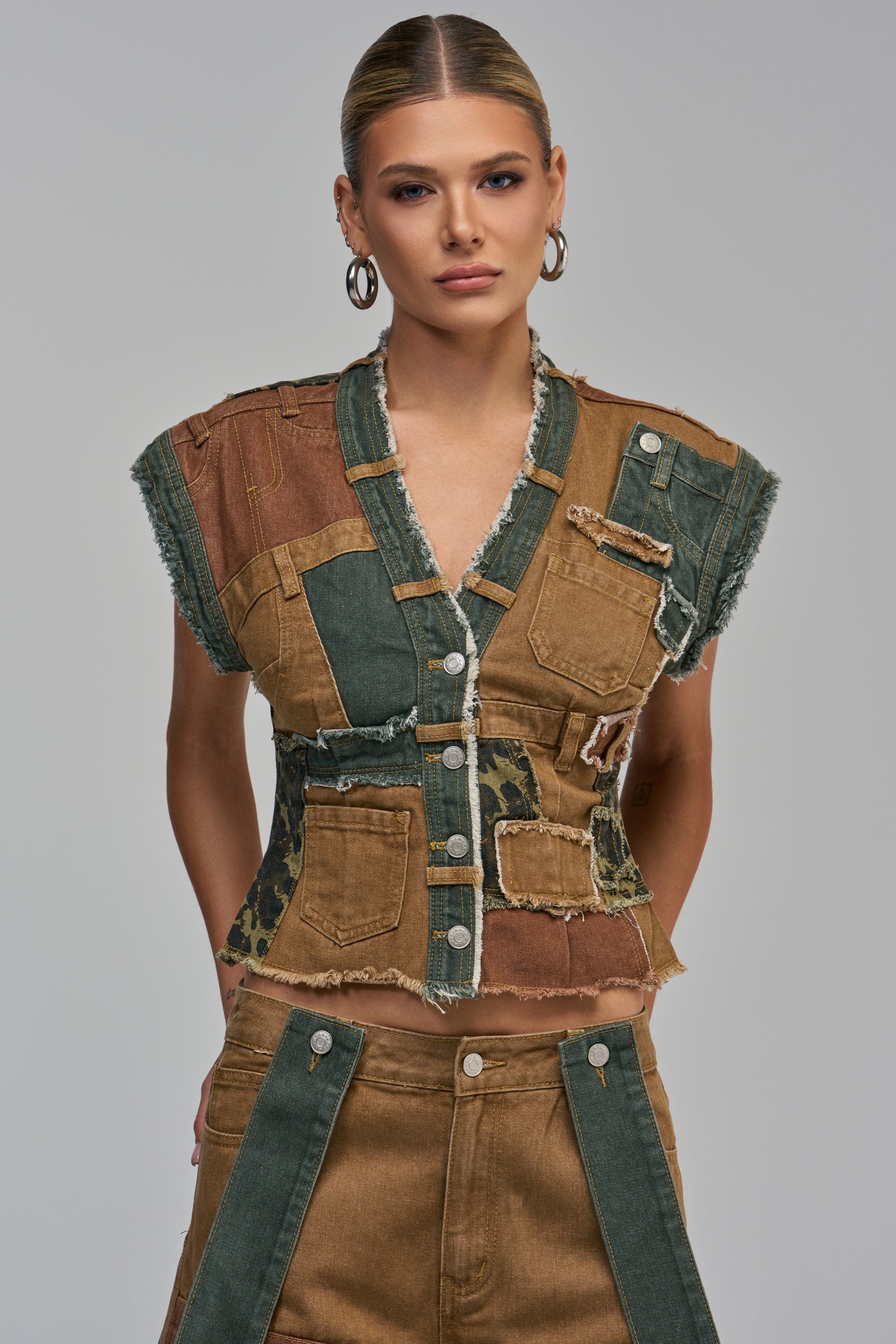 A model wears the LAYLA PATCHWORK DENIM BUTTON DOWN vest in shades of brown, olive, and black with frayed edges, paired with matching patchwork denim pants in a streetwear style.