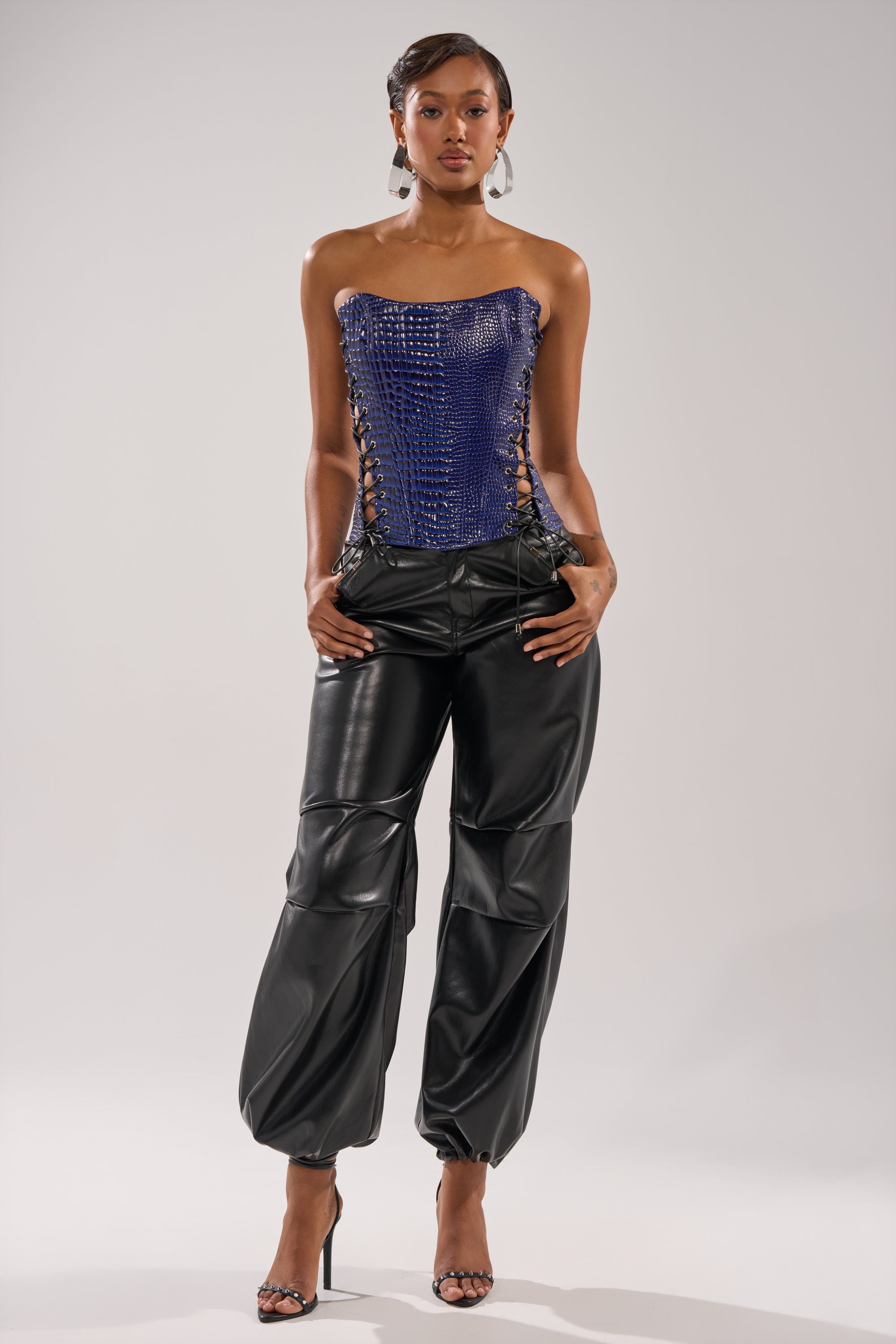 A woman poses confidently in black leather pants and the HIT THE SWITCH EMBOSSED CORSET with side lacing. She wears high-heeled sandals, drop earrings, and has short, styled hair against a plain background.