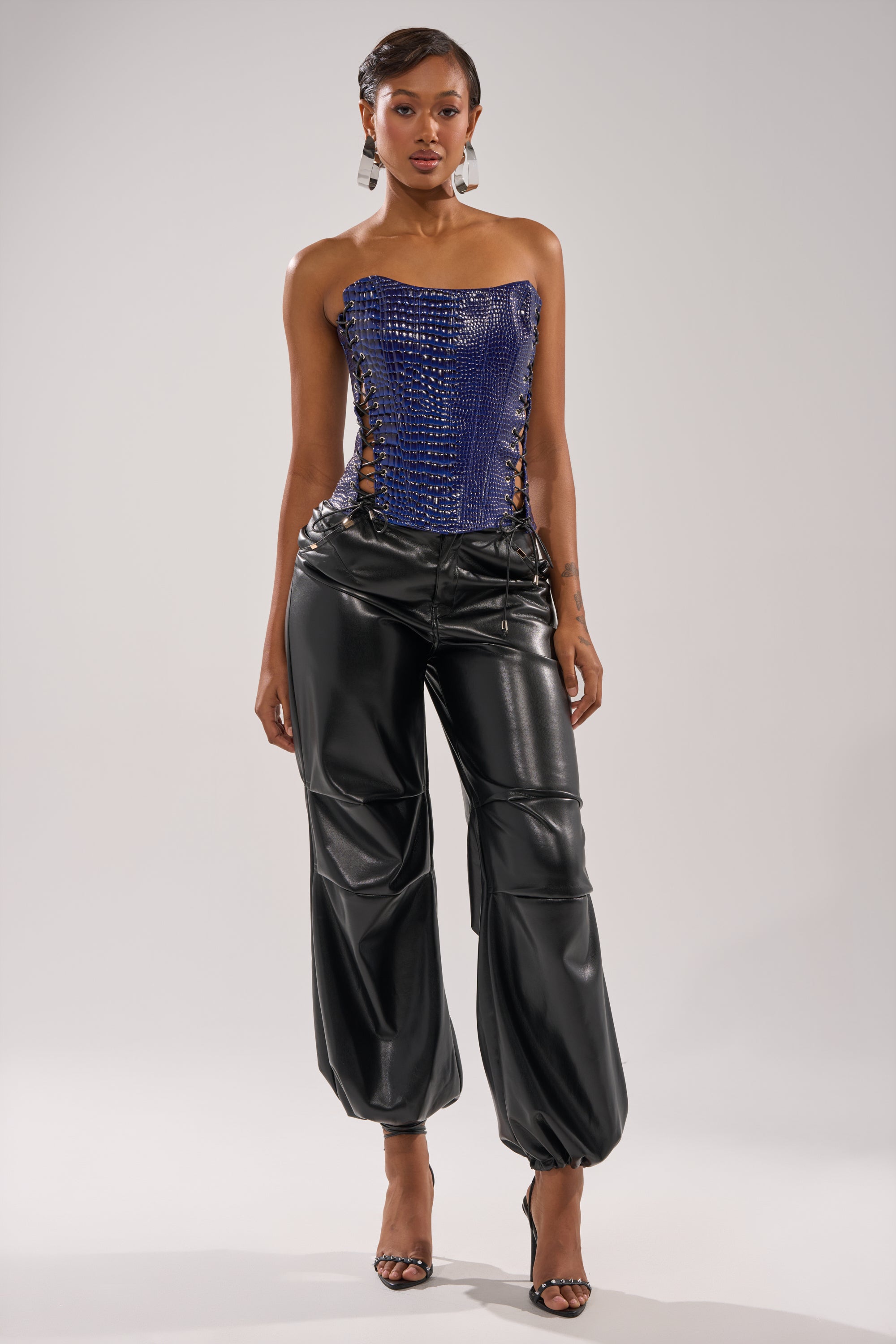 A woman faces forward wearing the HIT THE SWITCH EMBOSSED CORSET, black shiny leather pants with elastic hems, and black high-heeled sandals against a plain light background.
