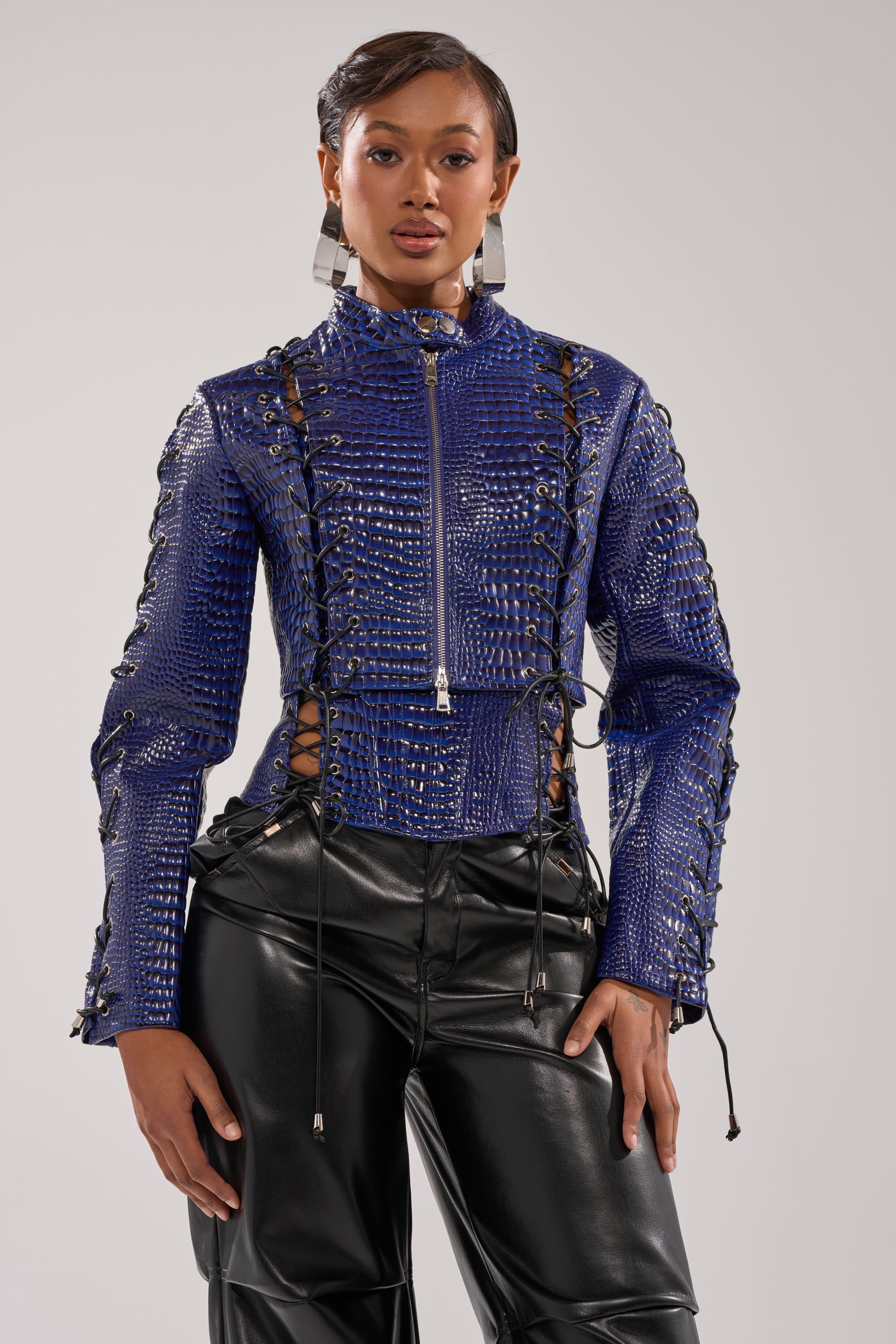 A woman stands confidently in the HIT THE SWITCH EMBOSSED MOTO, a shiny blue crocodile print zip-up jacket with lace-up details, paired with black faux leather pants and bold silver earrings, set against a plain light background. 1
