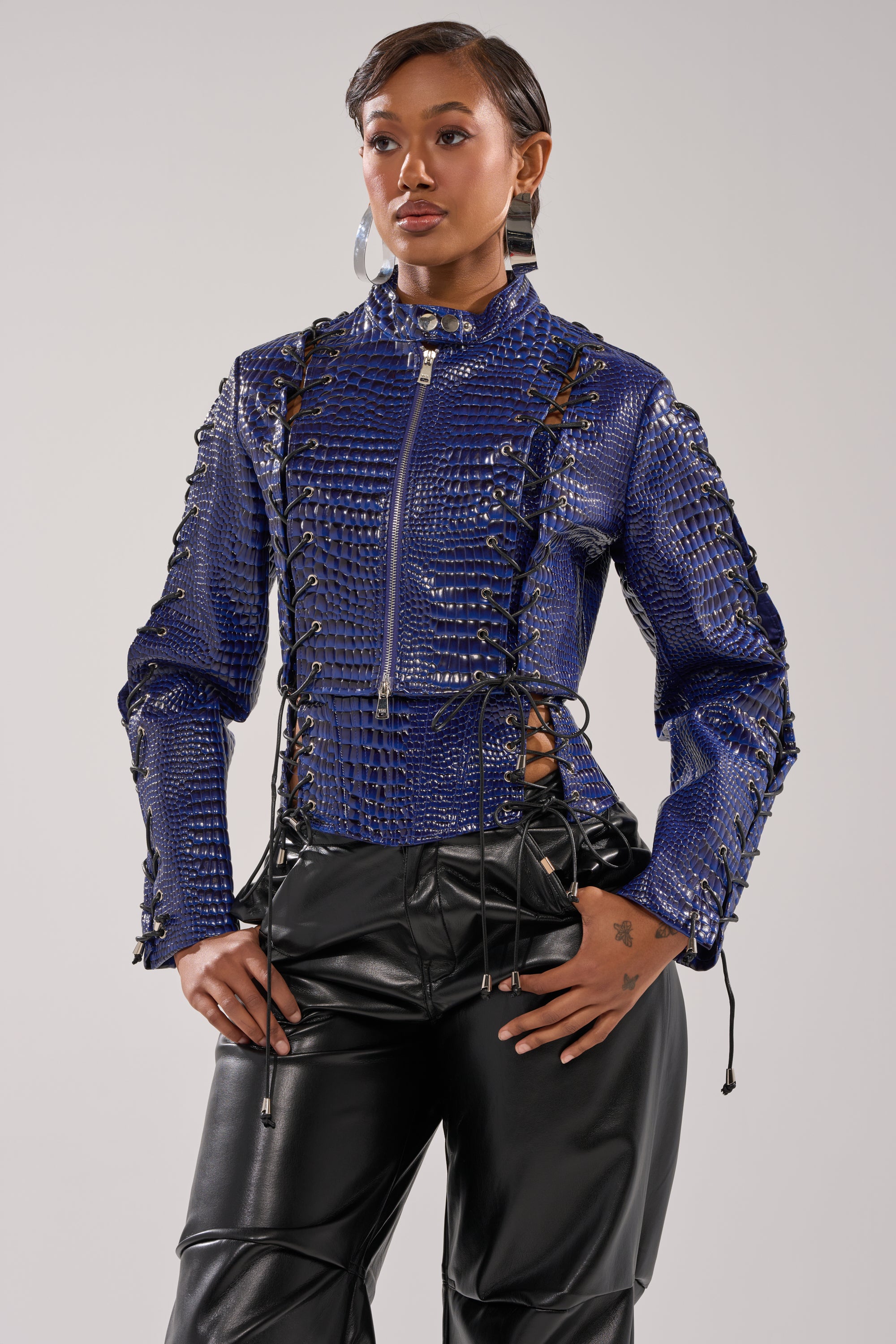 A model confidently wears the HIT THE SWITCH EMBOSSED MOTO—a shiny blue, cropped moto jacket with an embossed crocodile print and front zipper—paired with black faux leather pants, set against a plain light gray background. 2
