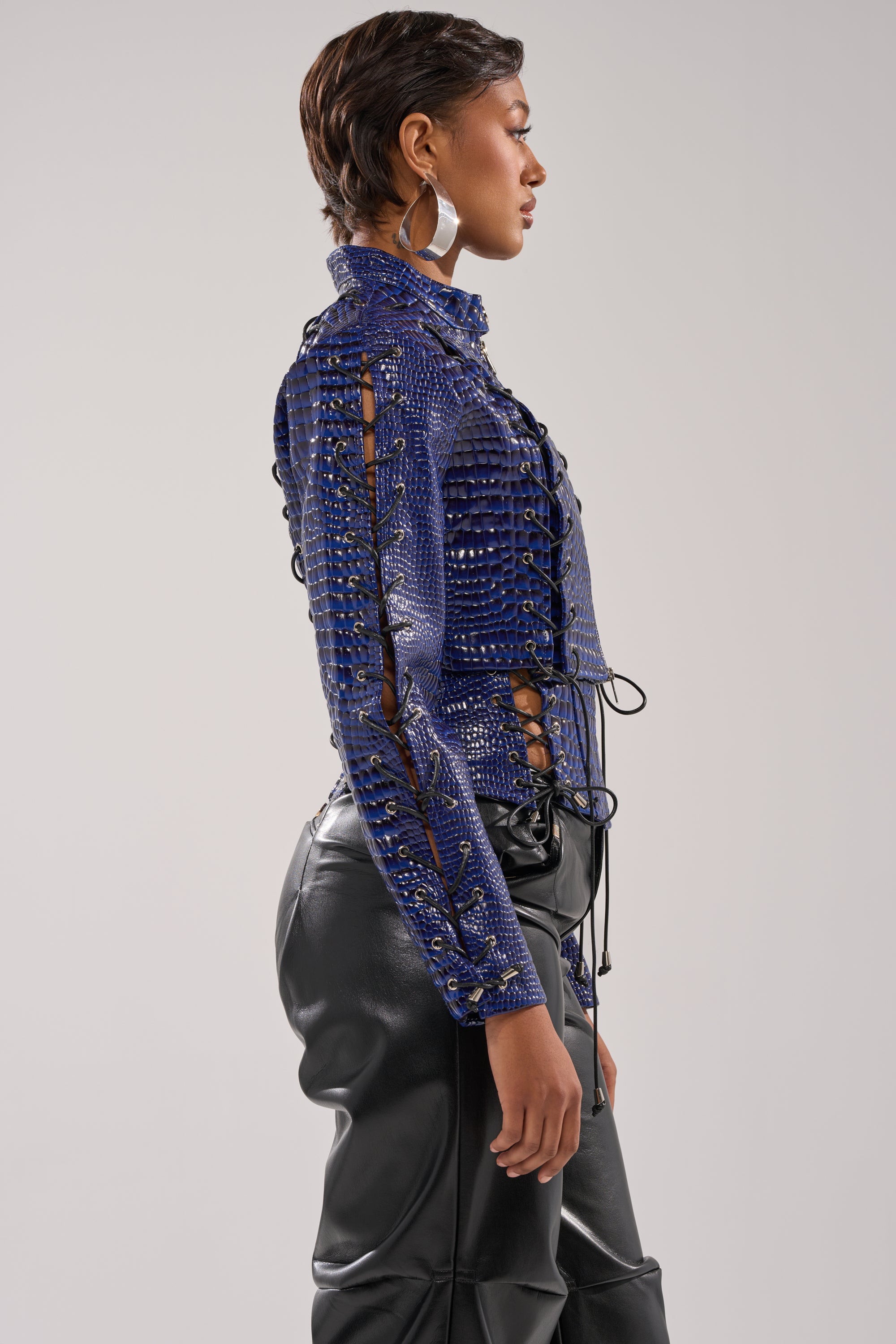 A woman in profile wears the HIT THE SWITCH EMBOSSED MOTO jacket, styled with shiny black leather pants, short hair, and large hoop earrings against a plain light background. 3