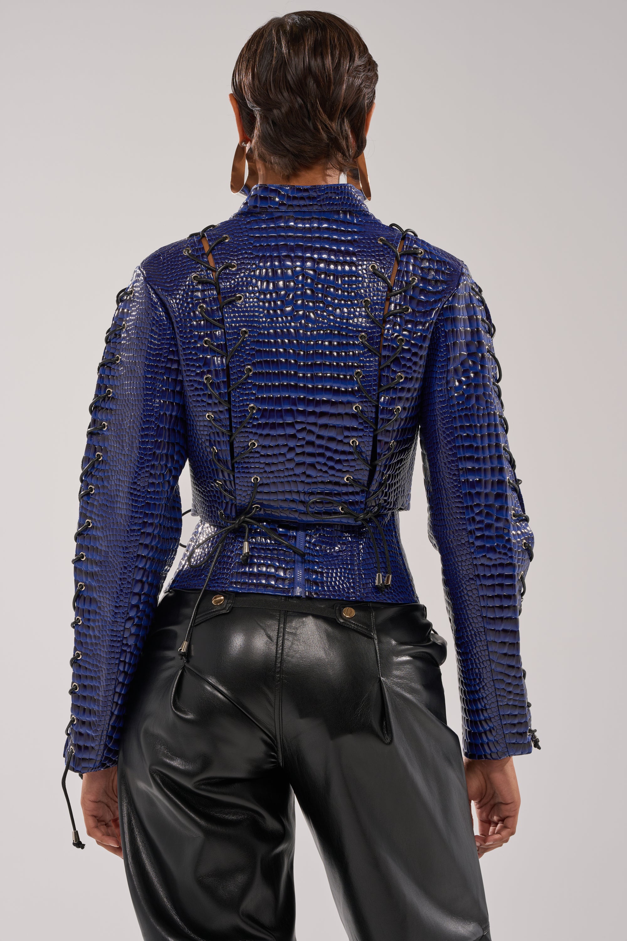 A person faces away, wearing the HIT THE SWITCH EMBOSSED MOTO blue faux patent leather jacket with crocodile print and black lace-up details, paired with high-waisted black leather pants against a plain light background. 4