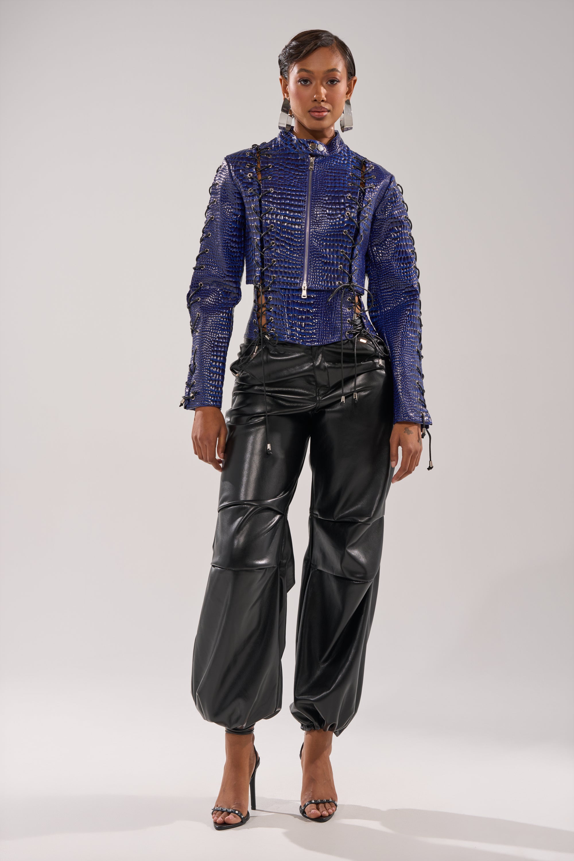 A woman stands confidently in the HIT THE SWITCH EMBOSSED MOTO, a shiny blue crocodile print jacket with a front zipper, paired with black leather cargo pants and strappy heels. Her neatly styled hair completes the chic look. 5