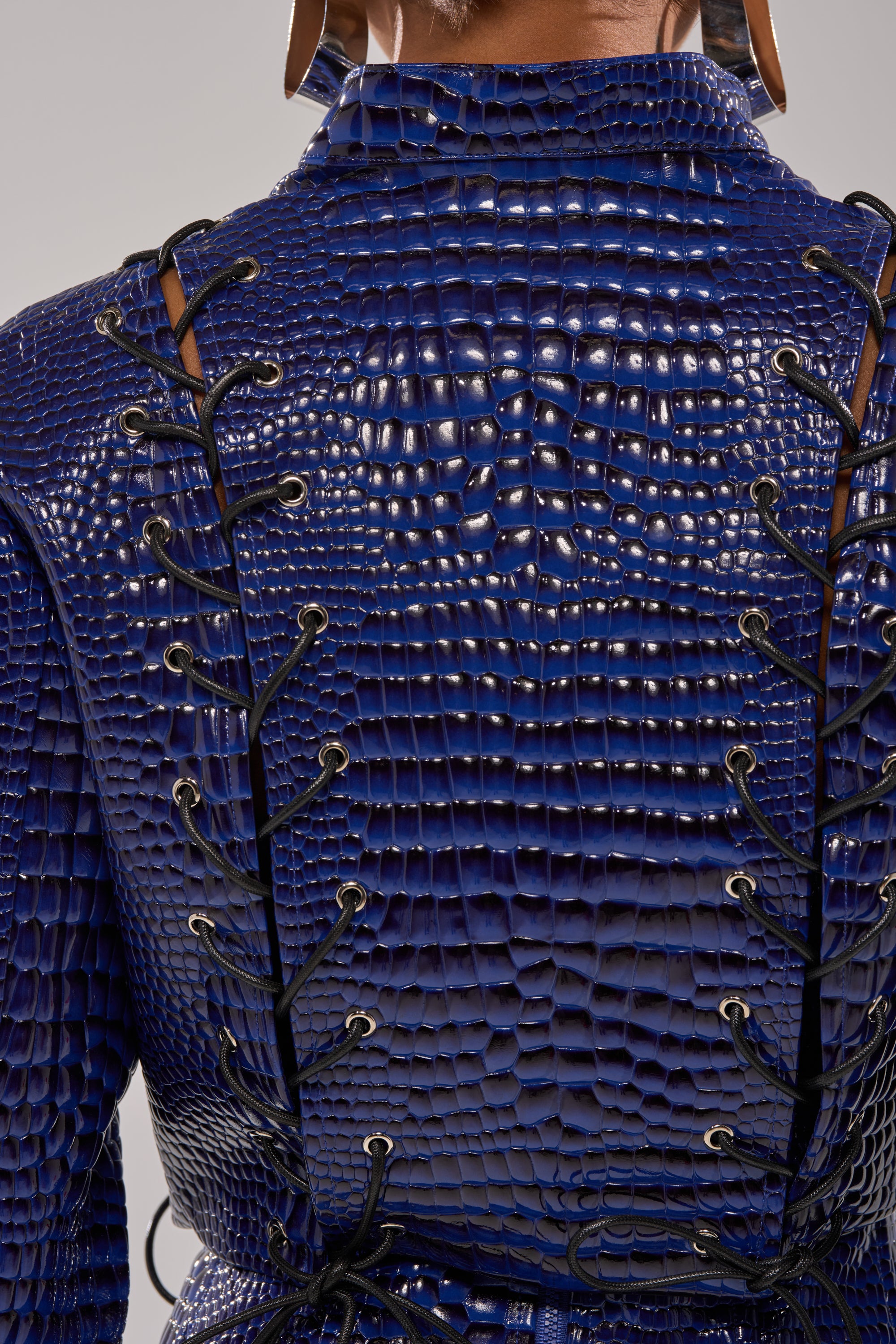 Close-up of the back of the HIT THE SWITCH EMBOSSED MOTO, a blue crocodile-print jacket featuring black laces crisscrossing through eyelets along the back and shoulders. 6