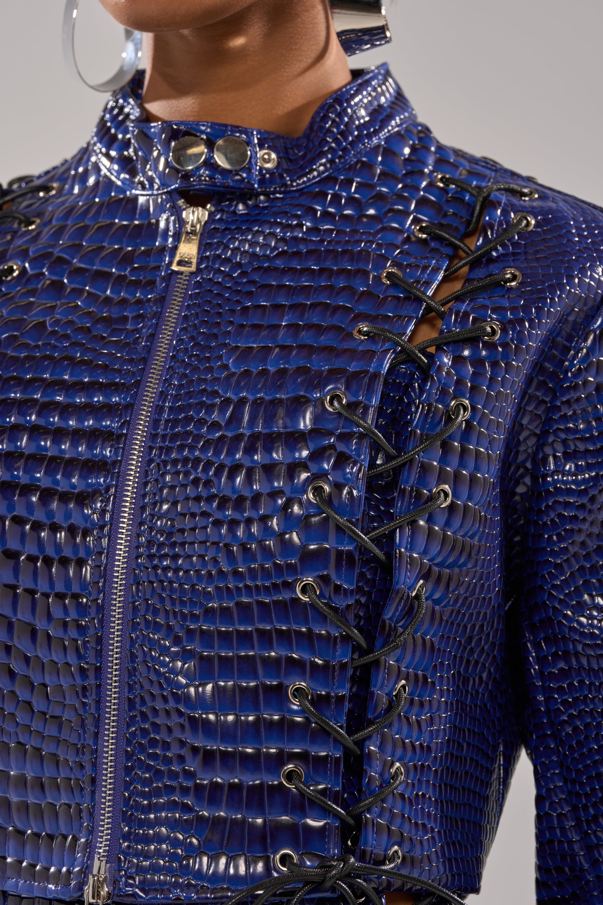Close-up of a person wearing the HIT THE SWITCH EMBOSSED MOTO jacket: shiny blue faux patent leather with crocodile embossing, zipper front, metallic snap buttons, and black lace-up details. 7