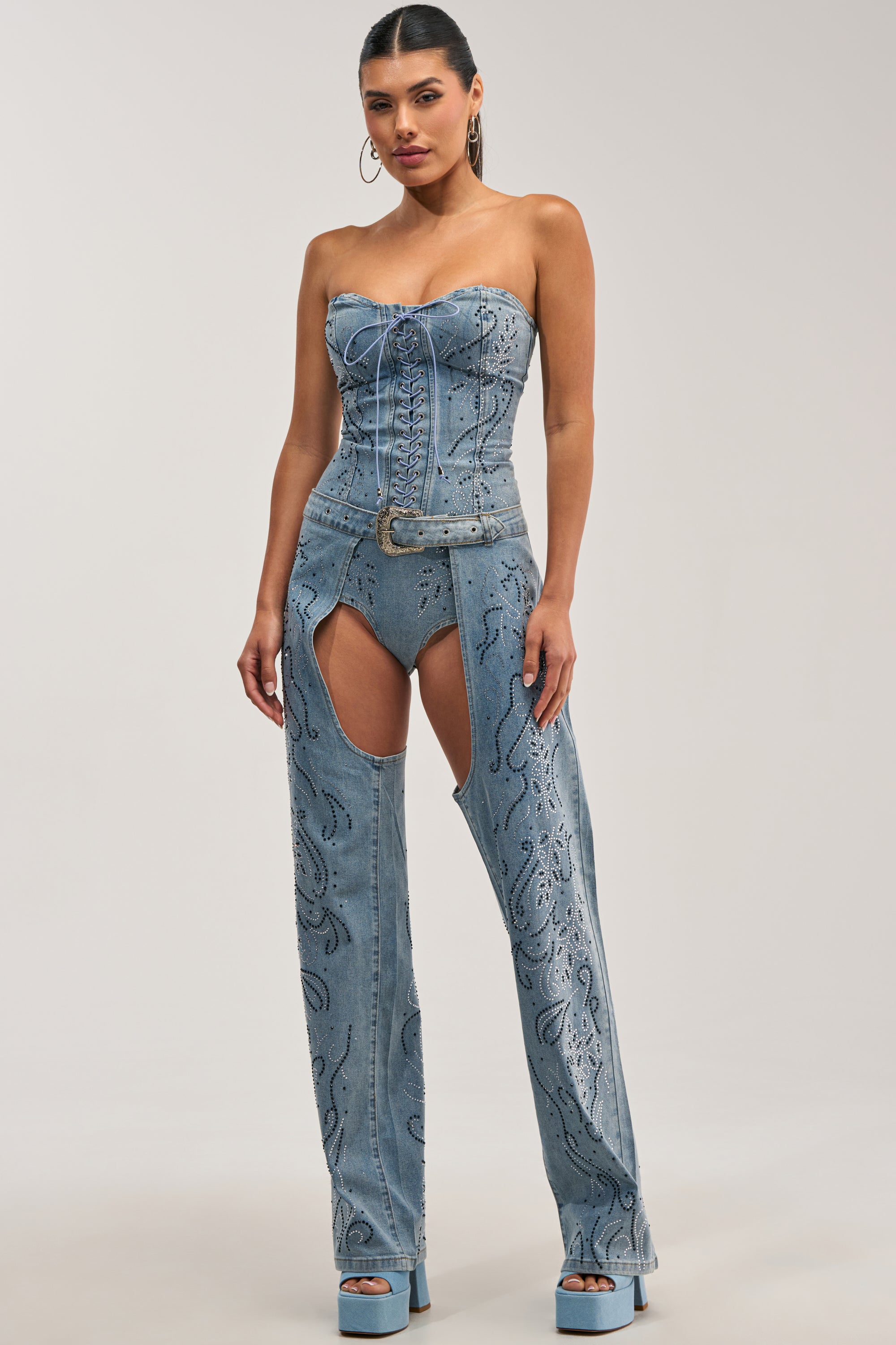 Dressed in the COWGIRL NIGHTS RHINESTONE DENIM CHAPS, a woman pairs high-waisted rhinestone-accented denim chaps with a strapless, lace-up denim corset and platform heels, capturing the essence of western festival fashion against a minimalist backdrop.